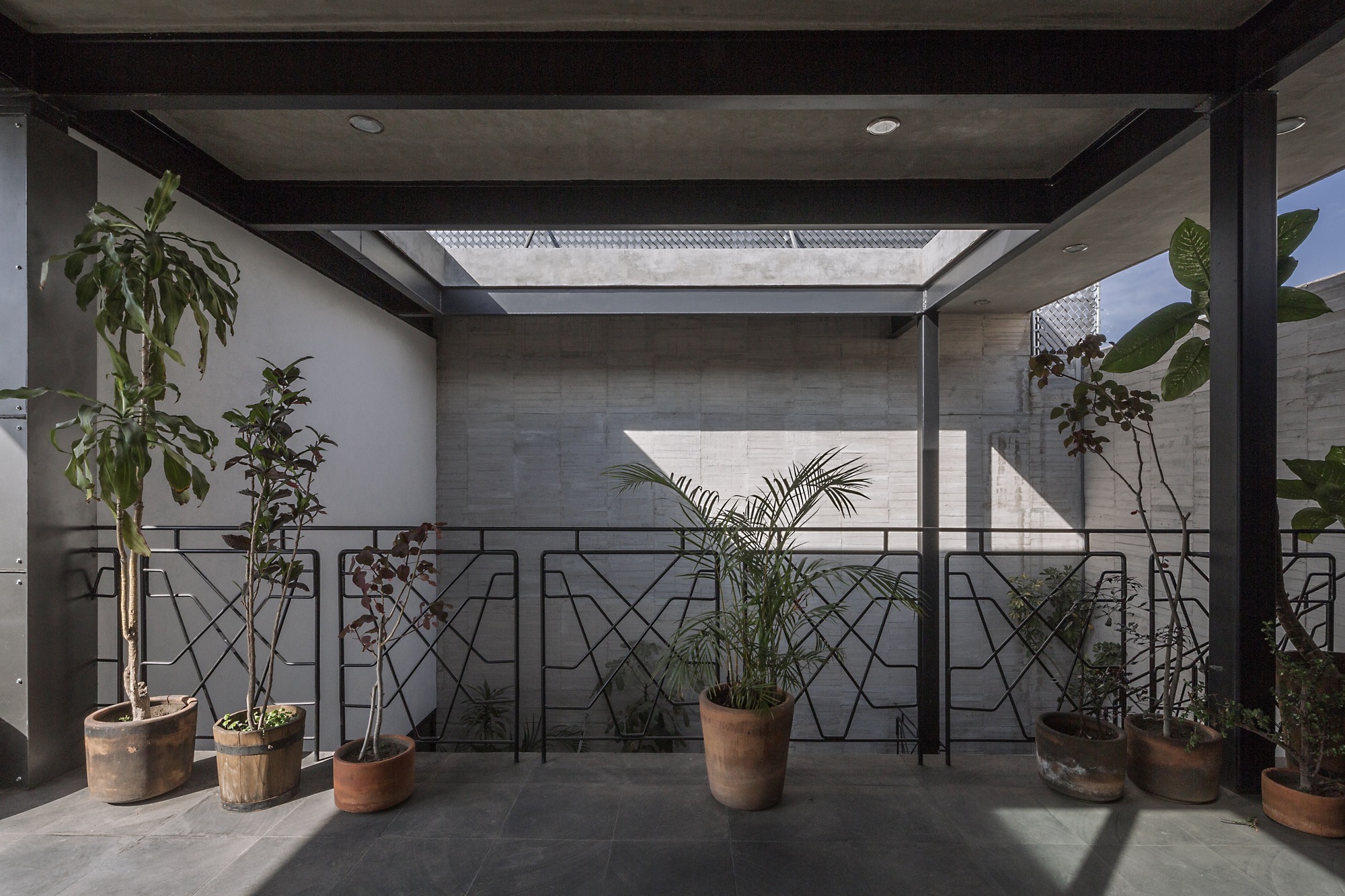 Covered upper terrace with steel beams, terracotta pots of plants and a geometric black railing in front of a board-marked concrete wall in sunlight