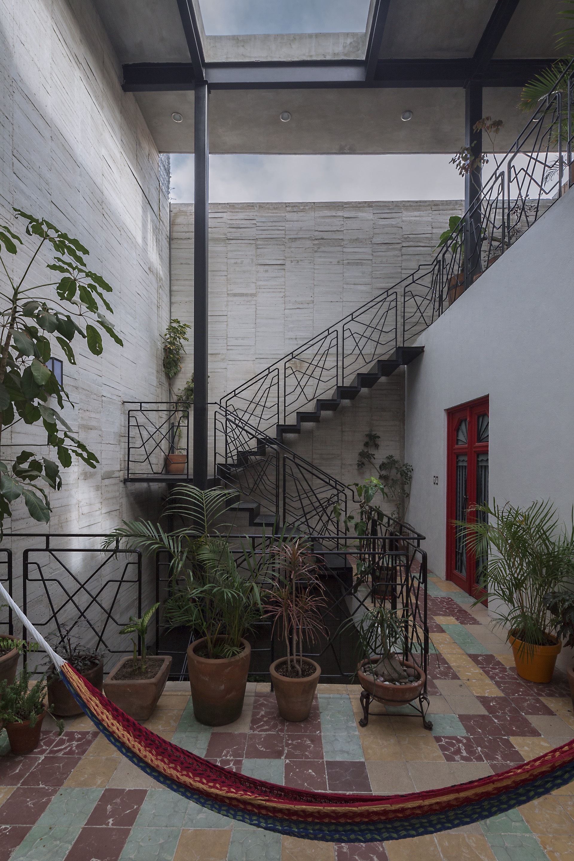 Courtyard with a black steel staircase and geometric railing beside a red door, a hammock hung over multicolored floor tiles