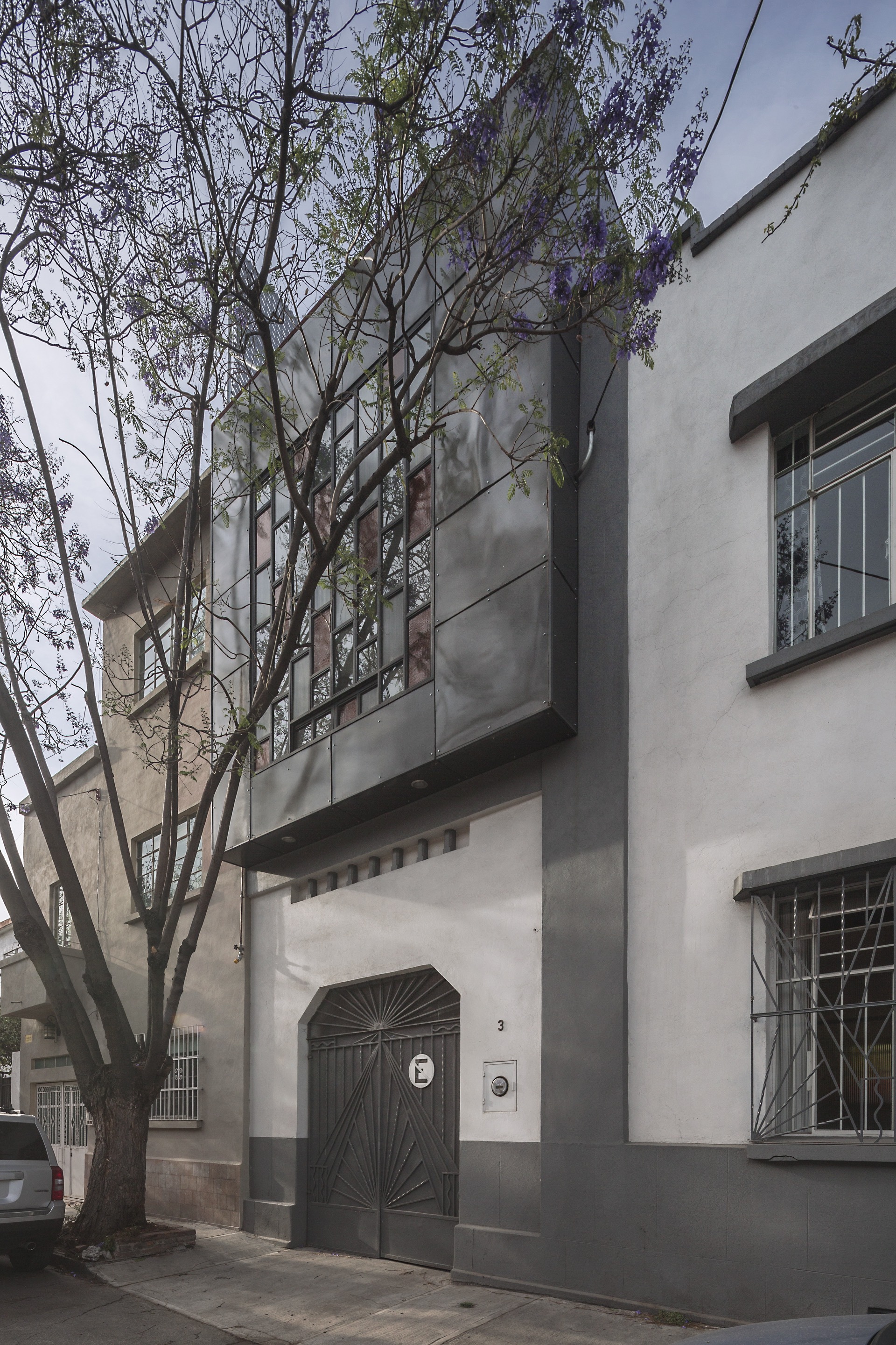 Street facade with a dark metal-clad bay of stained glass above an arched wooden gate, a flowering jacaranda in front