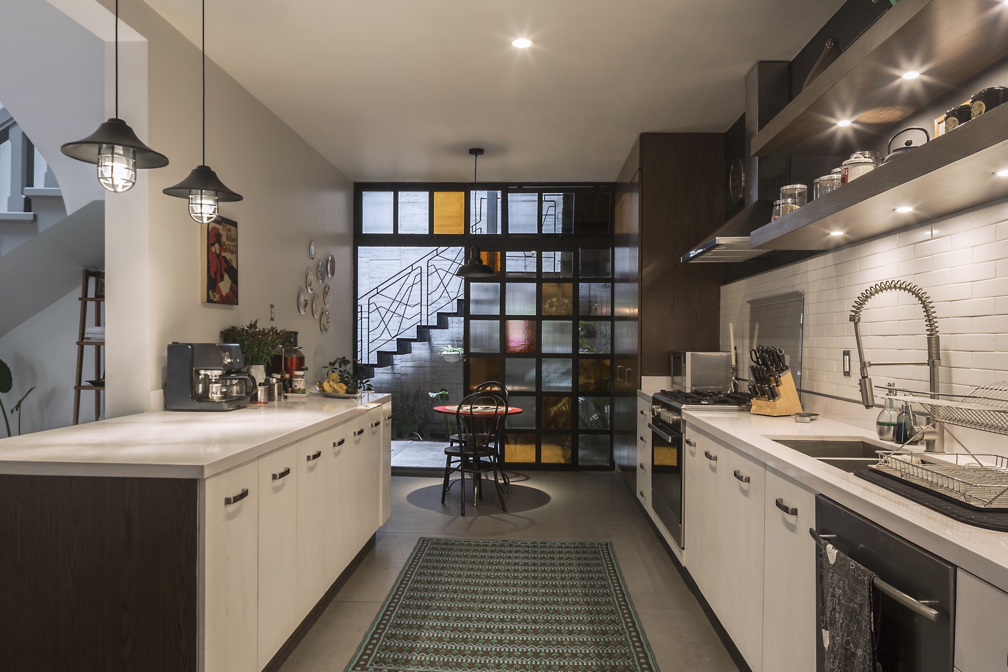 Galley kitchen with white cabinets and pendant lamps, ending in a stained glass wall that frames the courtyard stair