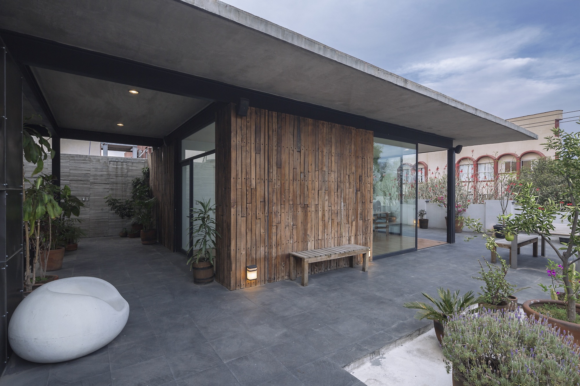 Covered rooftop terrace with a thin concrete roof slab, reclaimed timber wall and potted plants under a cloudy dusk sky