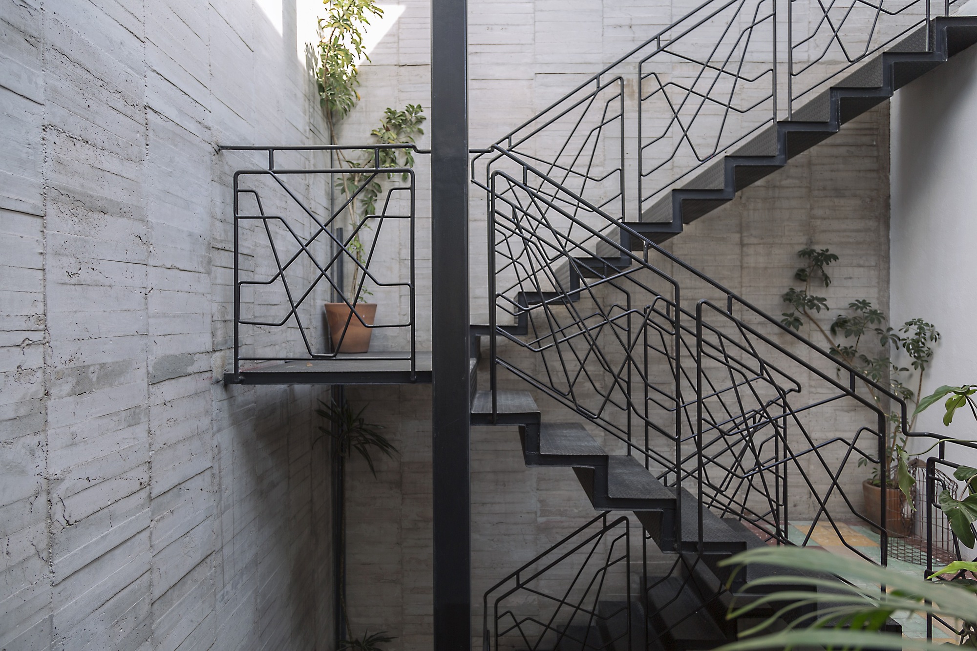 Black steel stair with geometric line-pattern railings rising past board-formed concrete walls and potted plants in a narrow light well