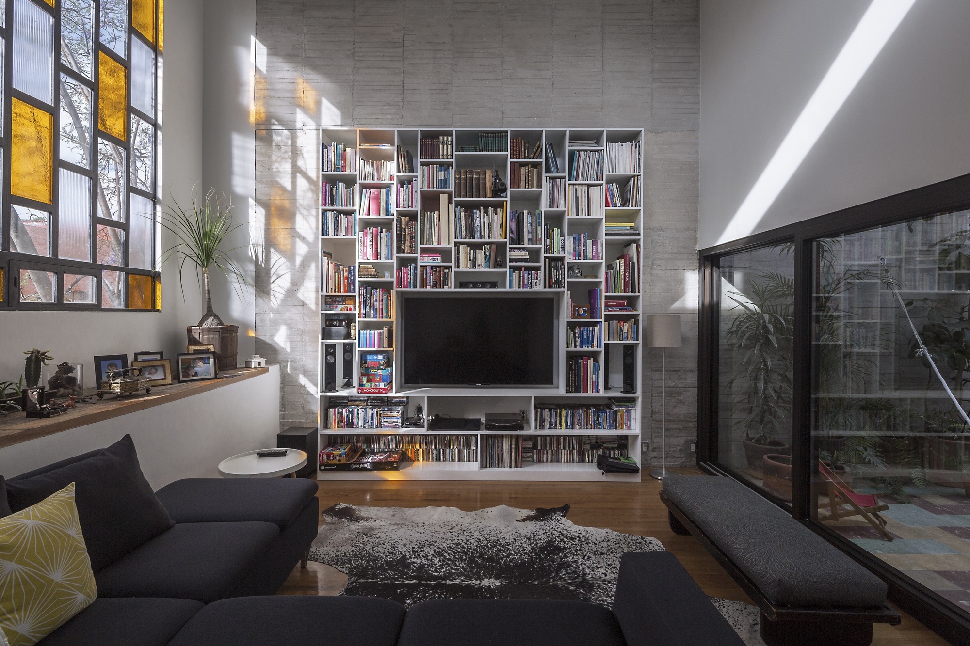 Living room with a full-height white bookshelf around a television, stained glass window and sunbeams crossing the concrete wall