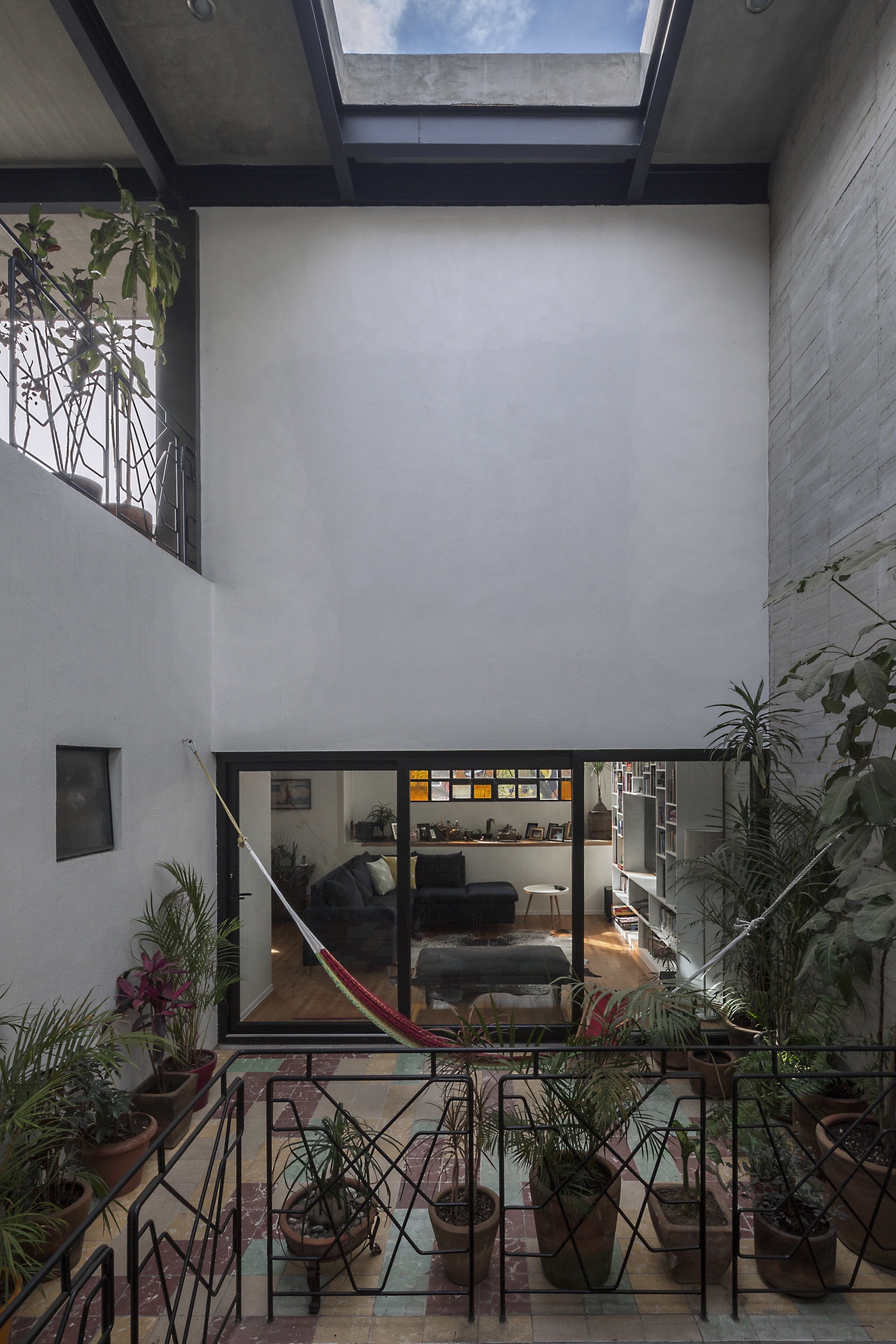 Double-height courtyard with a skylight, patterned metal railings, hanging hammock and potted plants in front of a glazed living room