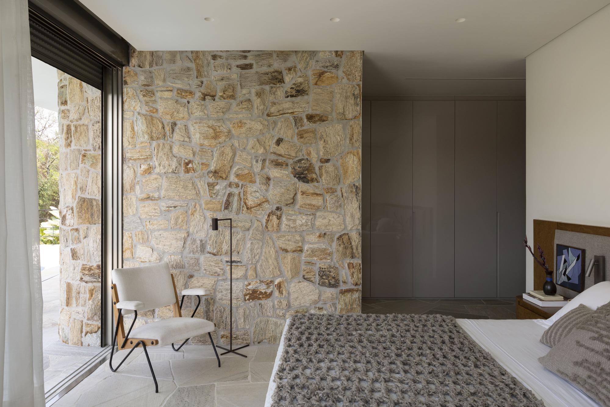 Bedroom with rough-cut stone wall adjacent to floor-to-ceiling glazing and a chair beside the window