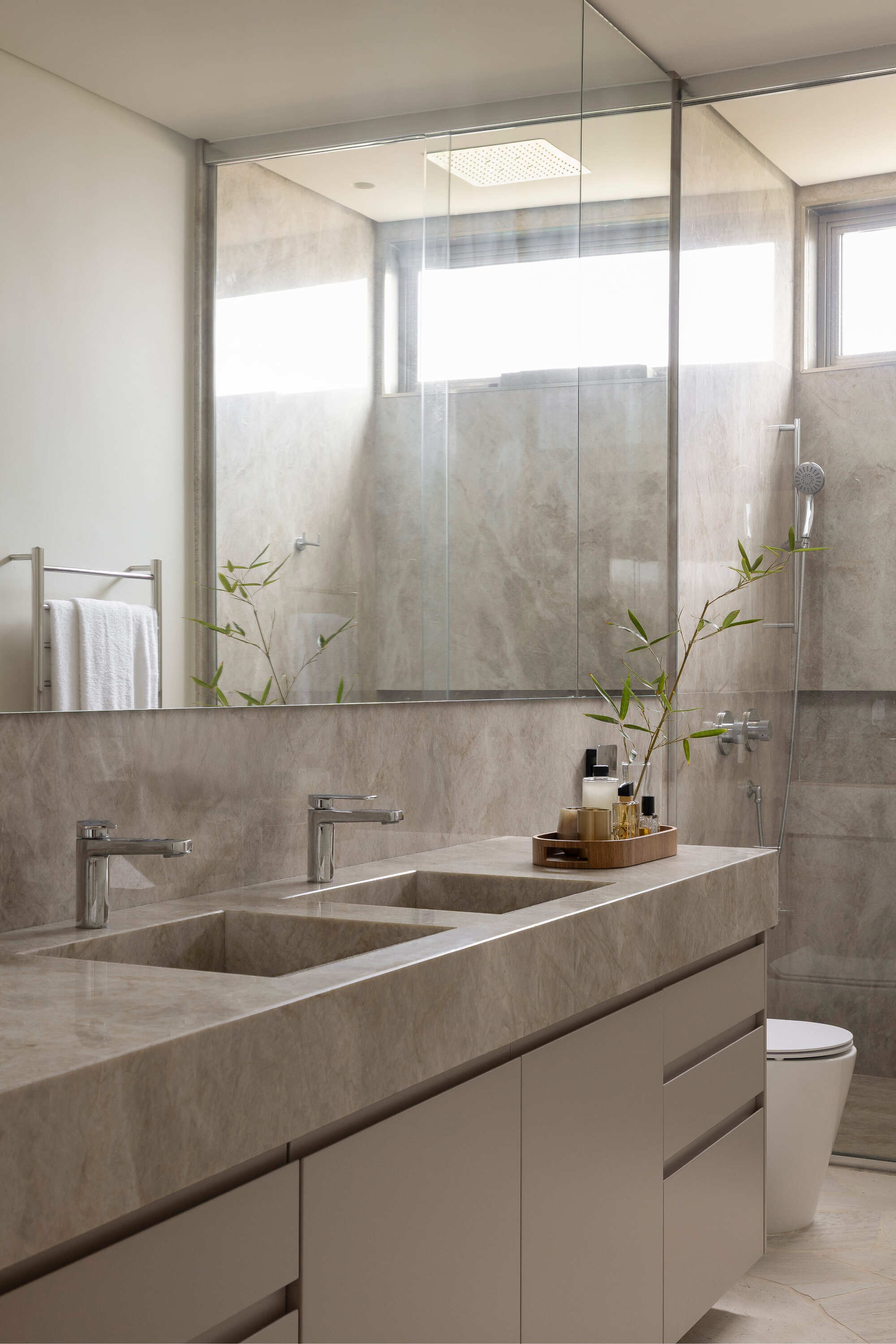 Bathroom vanity with grey stone countertop, twin sinks, and mirror reflecting glass shower enclosure with greenery