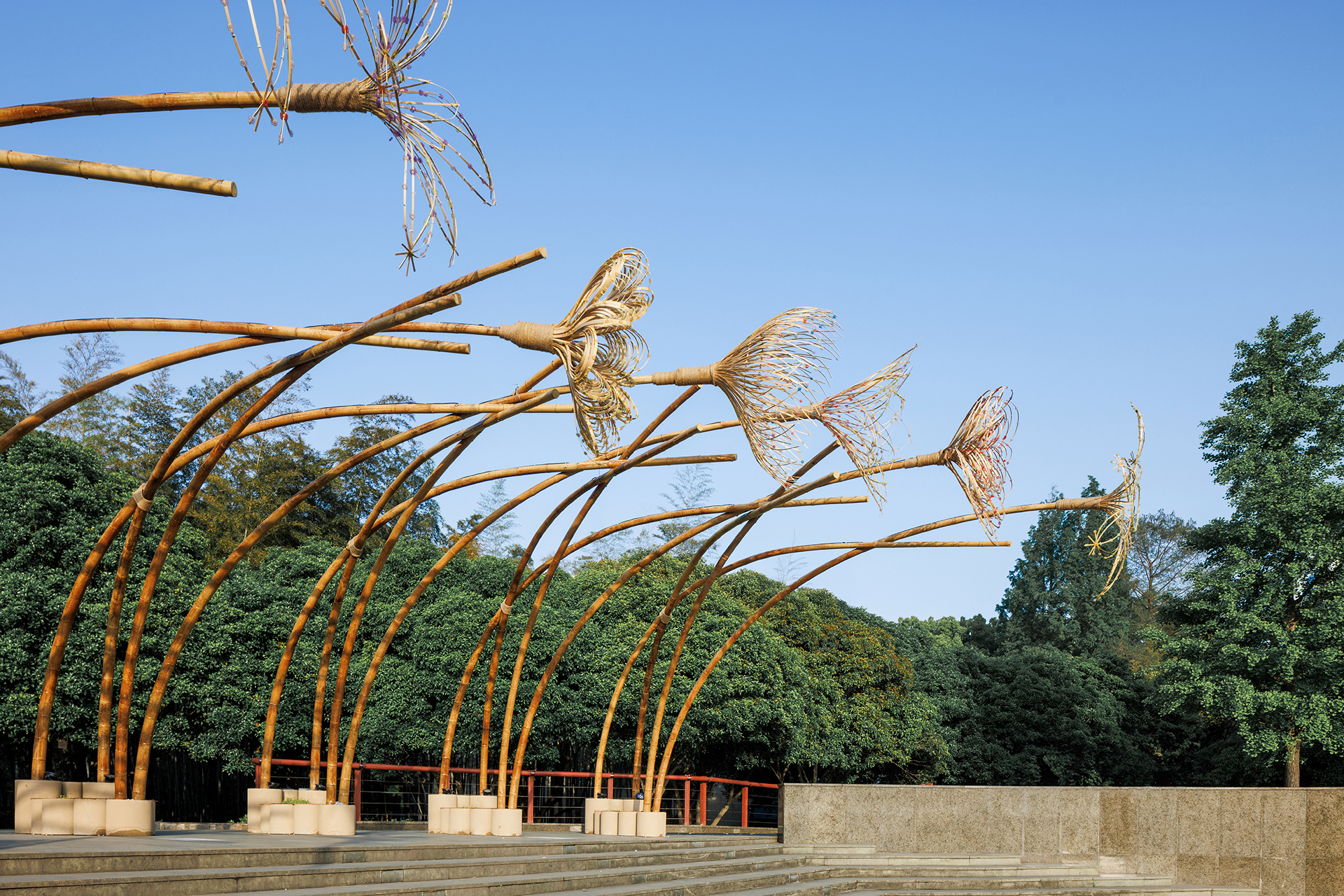 Curved bamboo arches with woven bird sculptures anchored to concrete footings under a clear blue sky