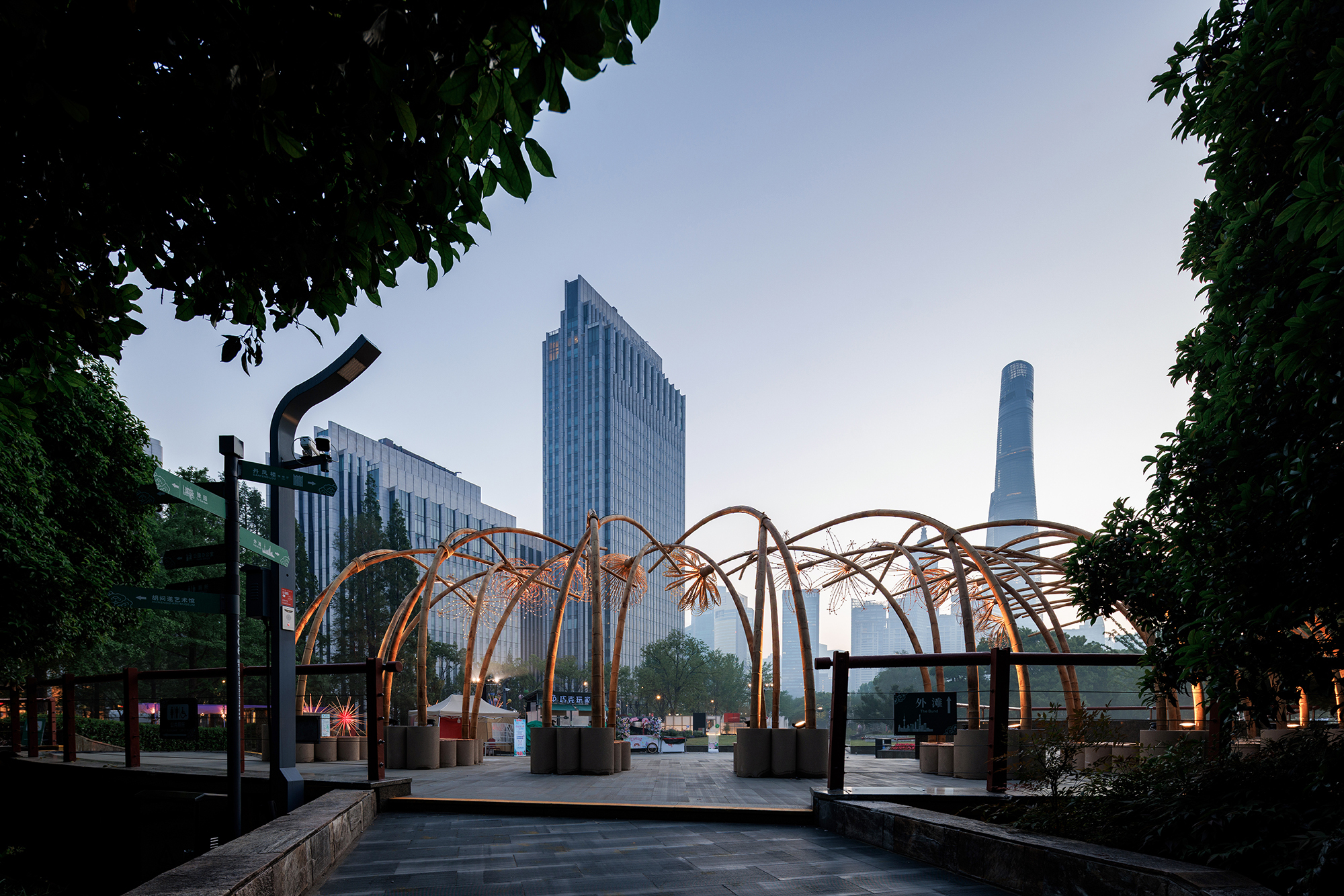 Illuminated bamboo canopy in a public plaza with highrise towers visible in the evening haze
