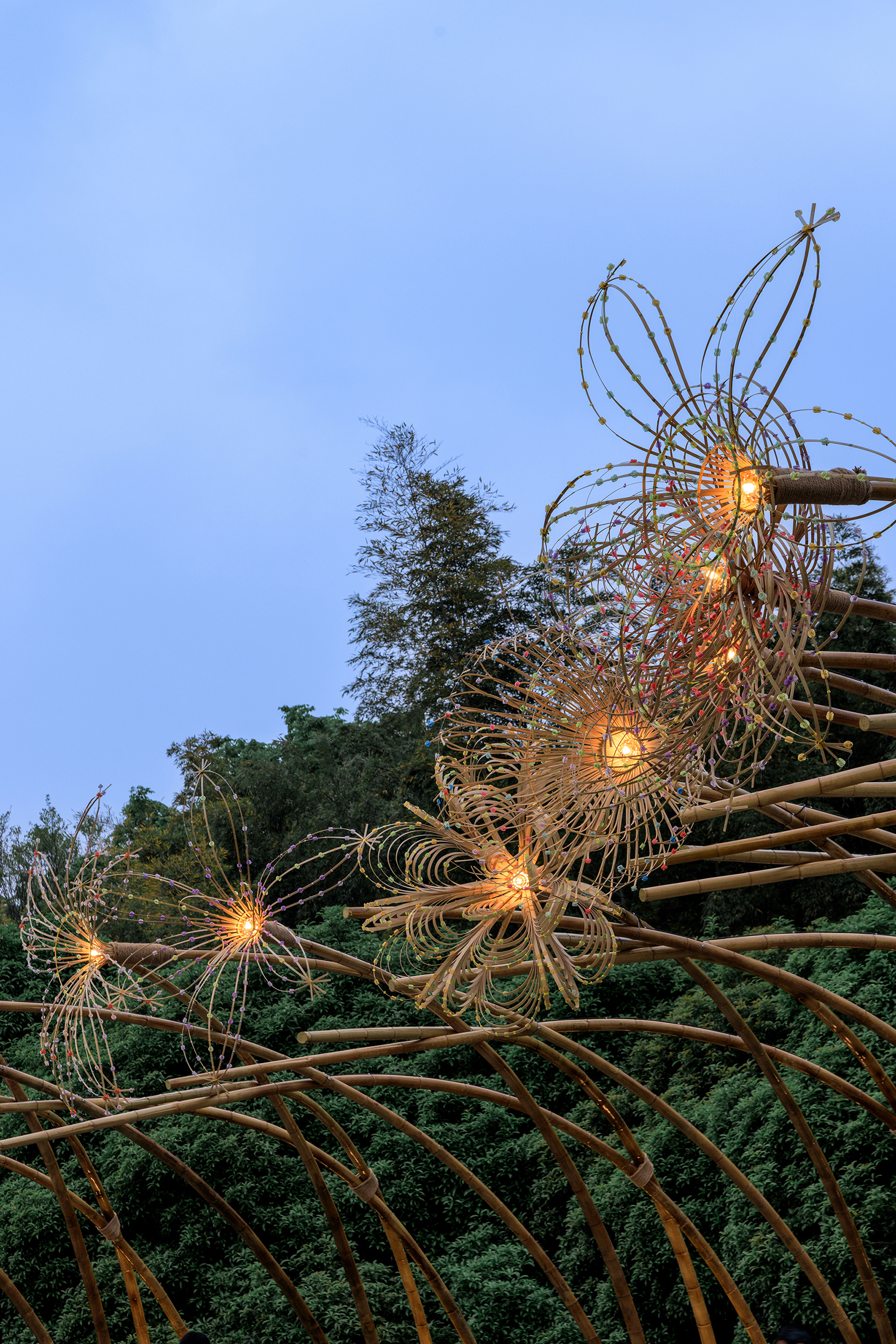 Bamboo sculpture with integrated lighting illuminating wire flower forms at dusk among surrounding trees