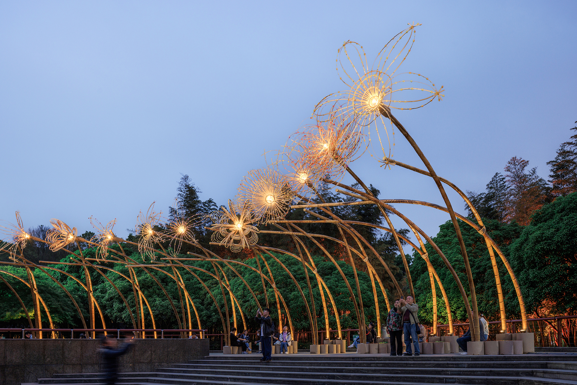 Arched steel structure with lit wire flowers and visitors walking beneath at evening