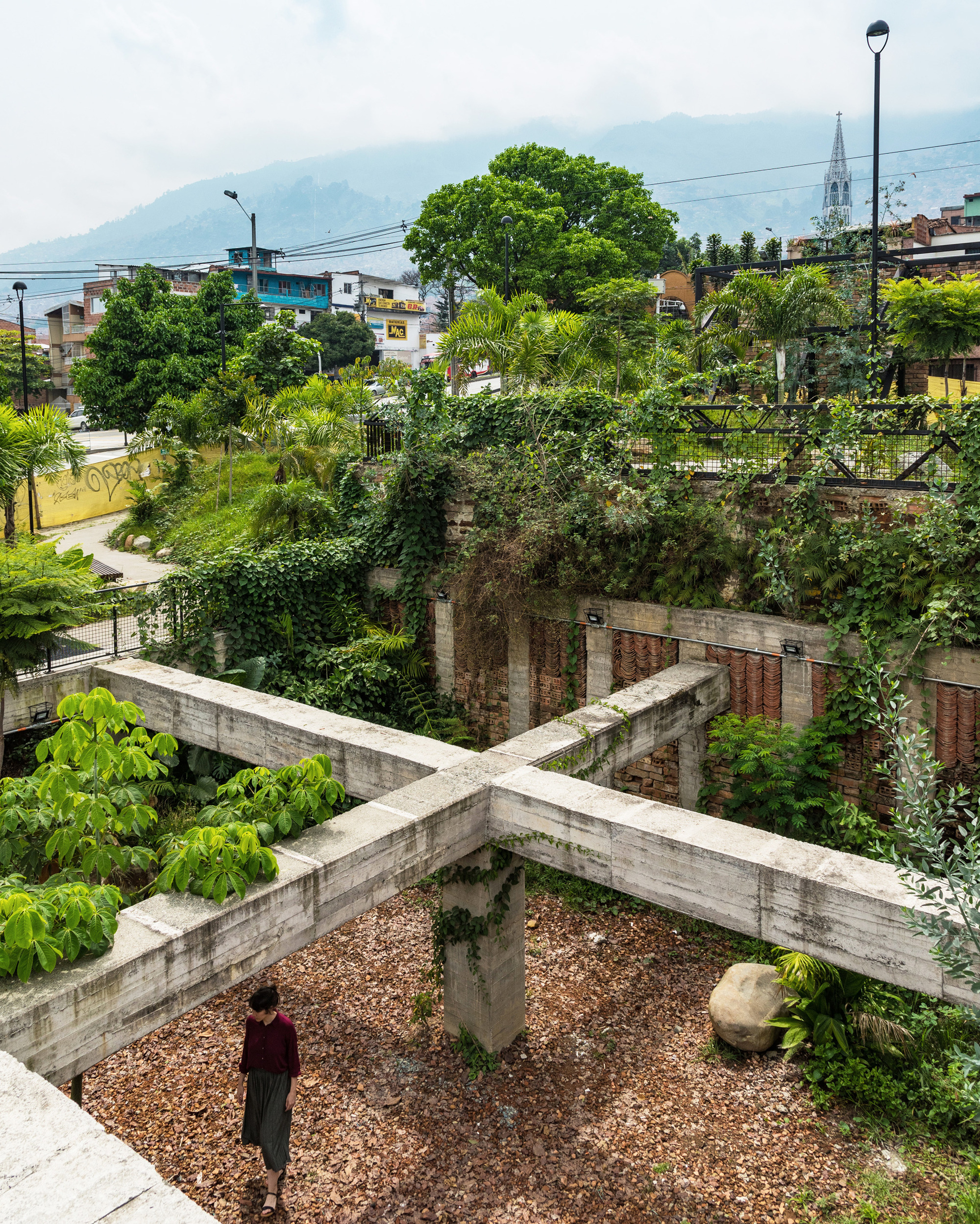Concrete crossbeam structure over gravel courtyard with ivy-covered walls and a person standing below