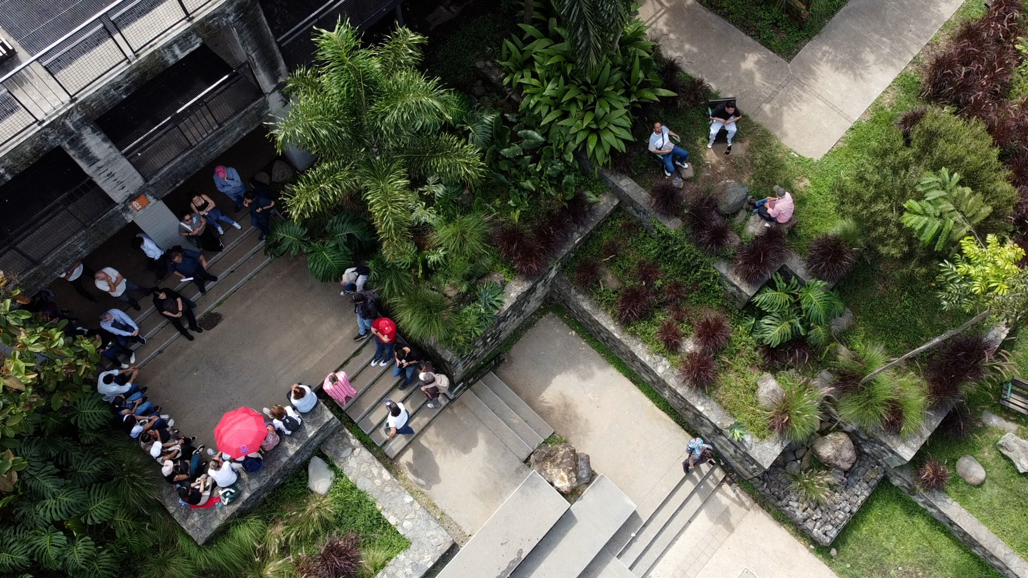 Aerial view of tiered concrete plaza with planted beds, visitors gathering, and a red umbrella