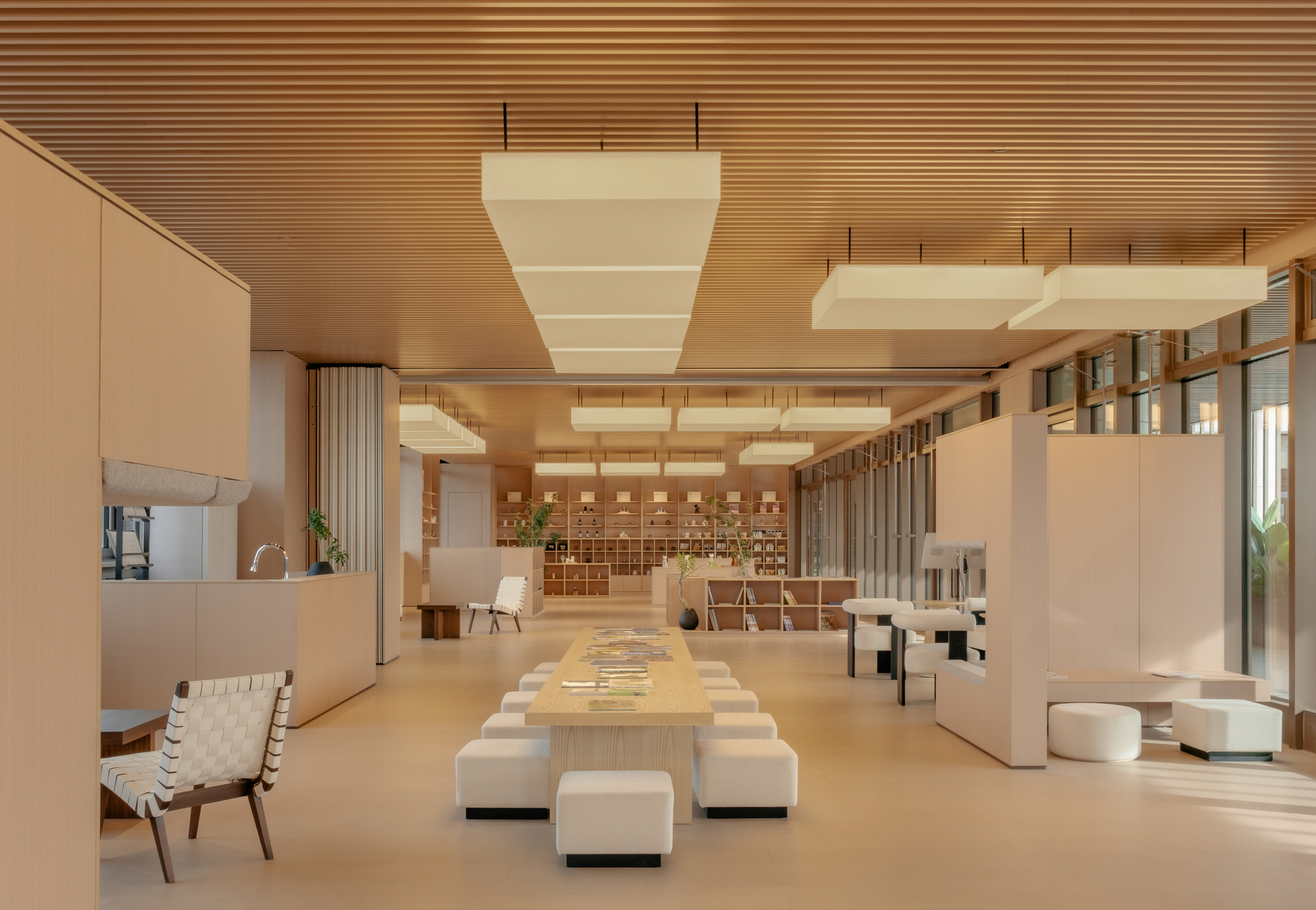 Open lounge with suspended baffles, long communal table, white seating cubes, and gridded shelving at rear