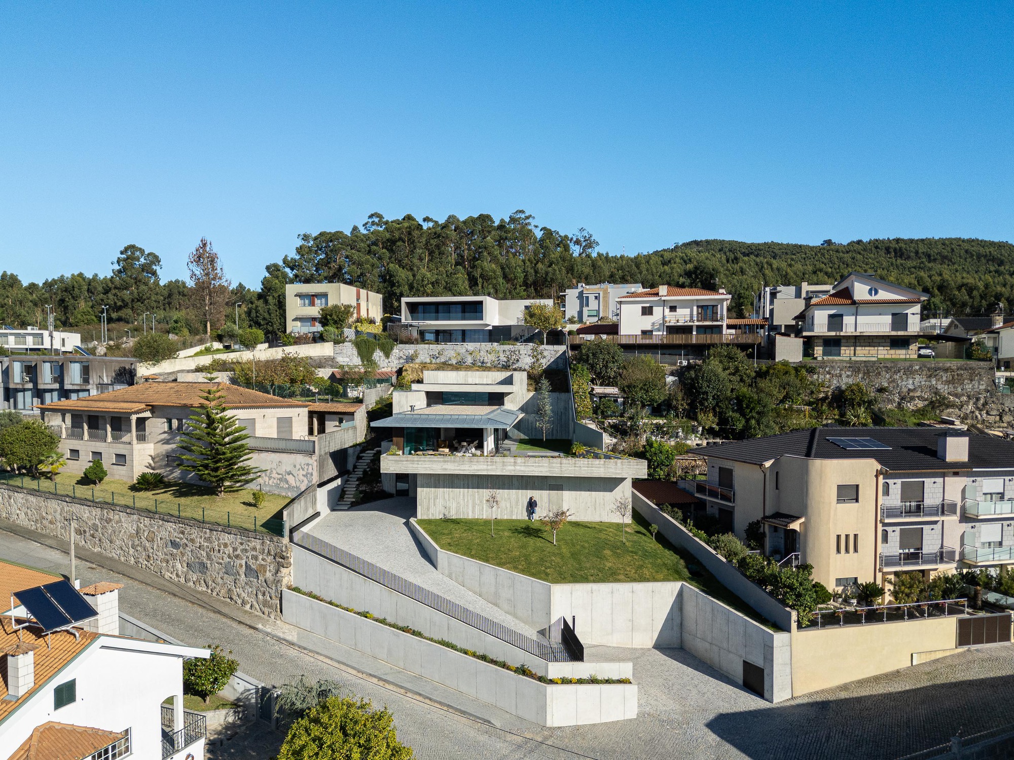 Aerial view of terraced concrete volumes stepping down a hillside among neighboring houses and trees