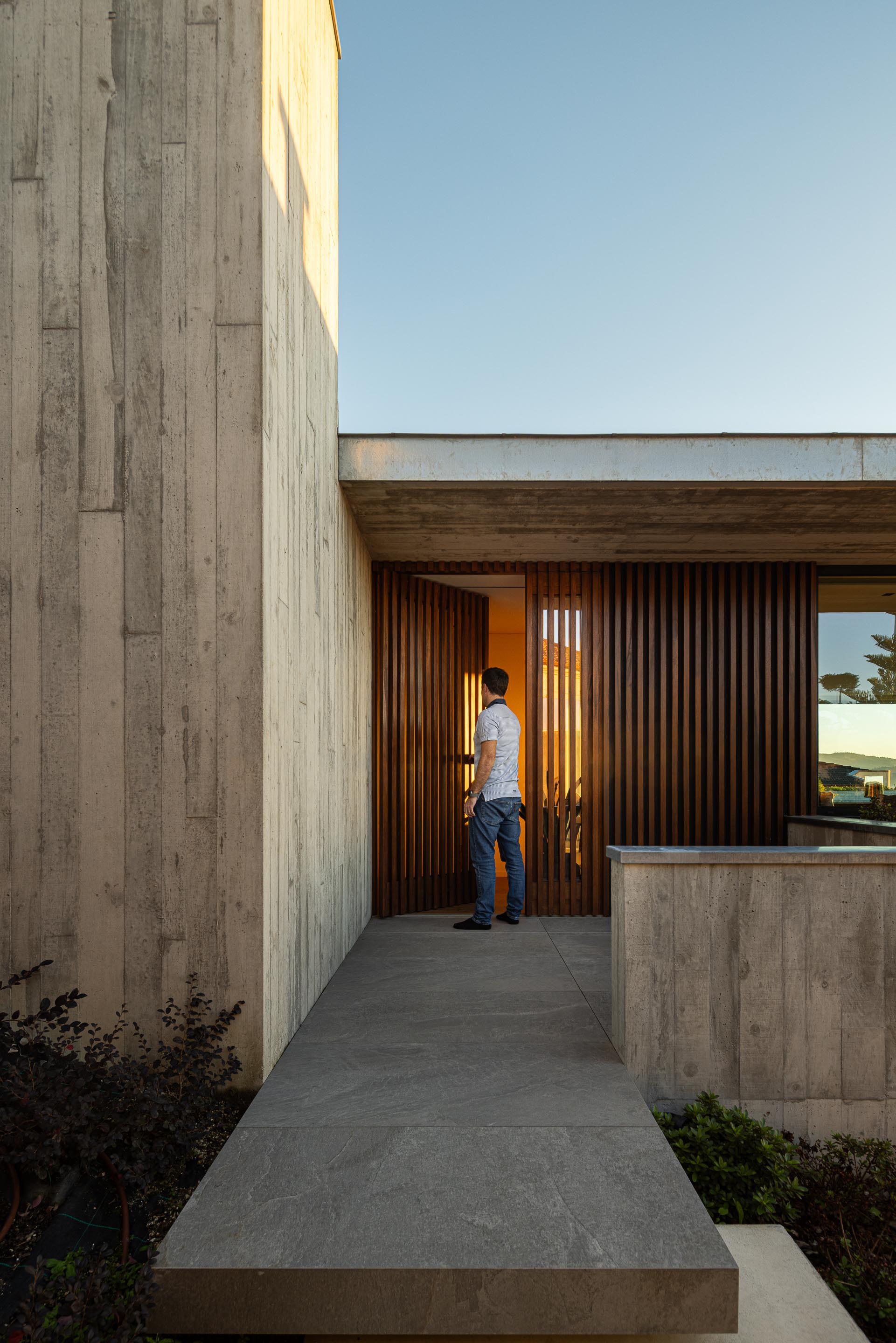 Entry passage with board-formed concrete walls and timber slatted screen as a figure approaches at dusk
