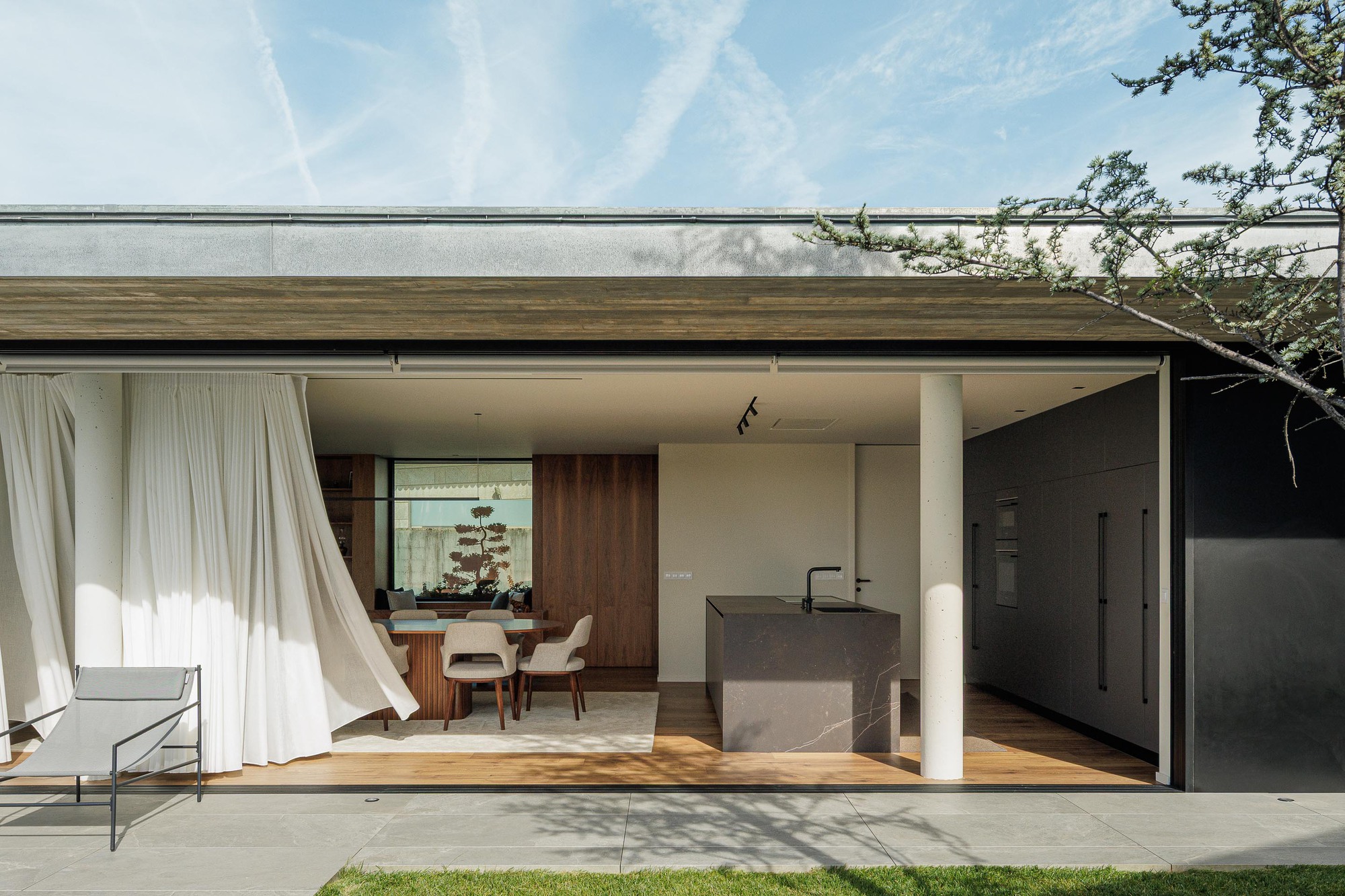 Covered outdoor terrace with flowing curtains opening to kitchen island and timber-clad dining area beyond