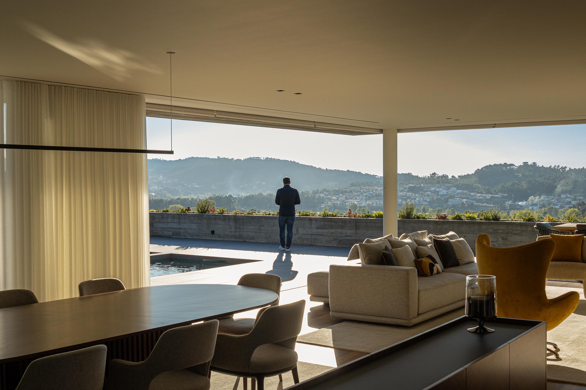 Open living area with a figure standing on the terrace overlooking distant hills at golden hour
