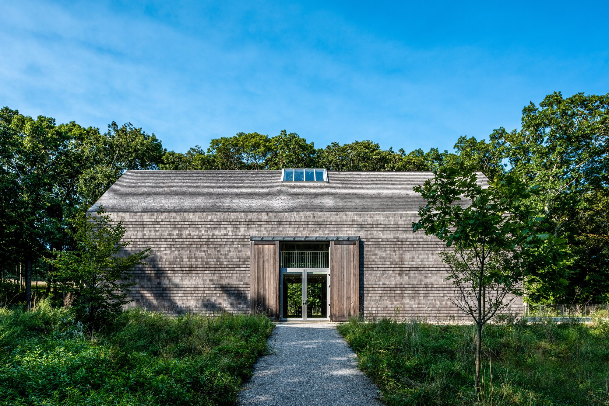 Cedar shingle facade with central glass entry door and roof monitor framed by dense oak trees