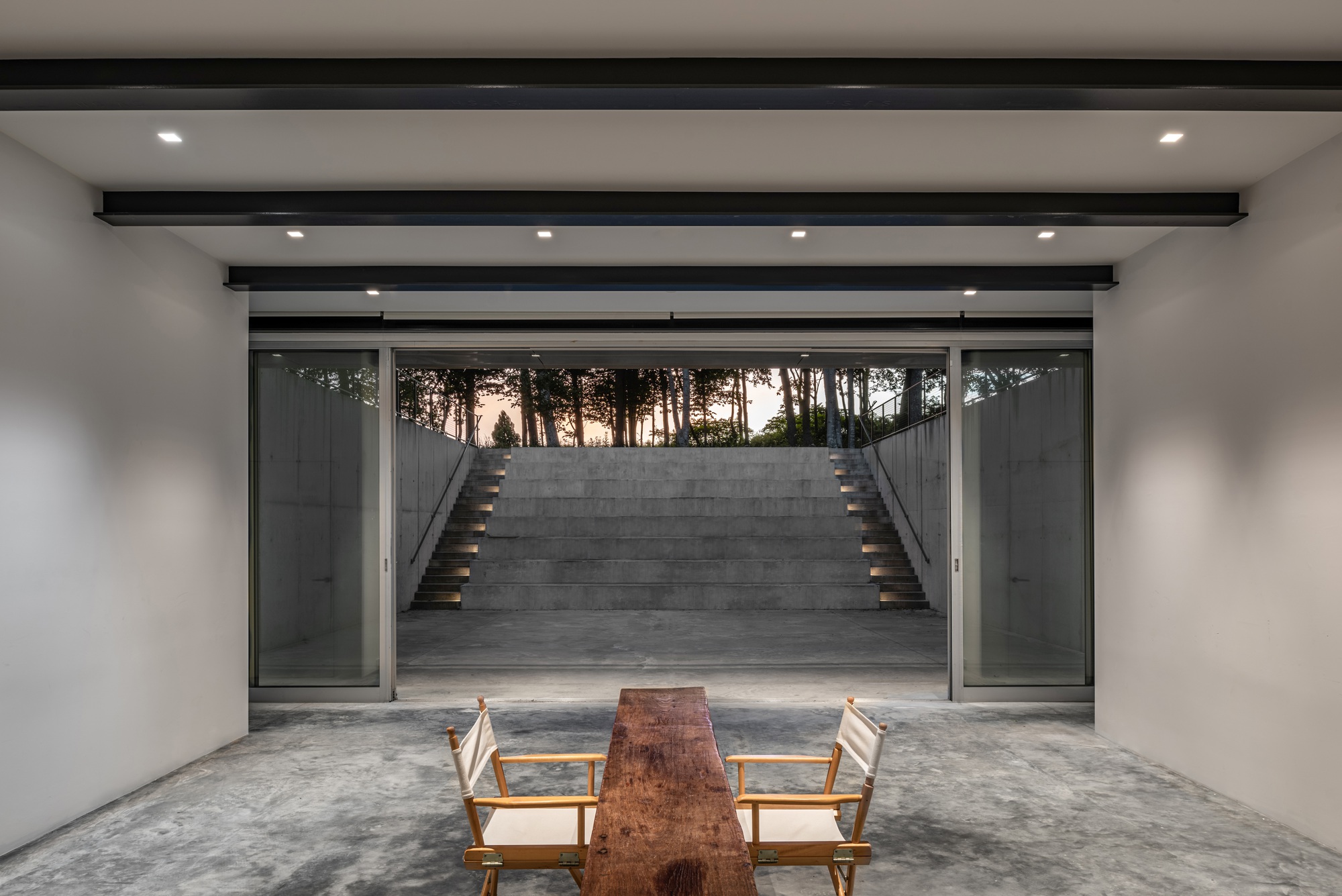 Interior room framing a concrete staircase ascending to trees at dusk through full-height glazing