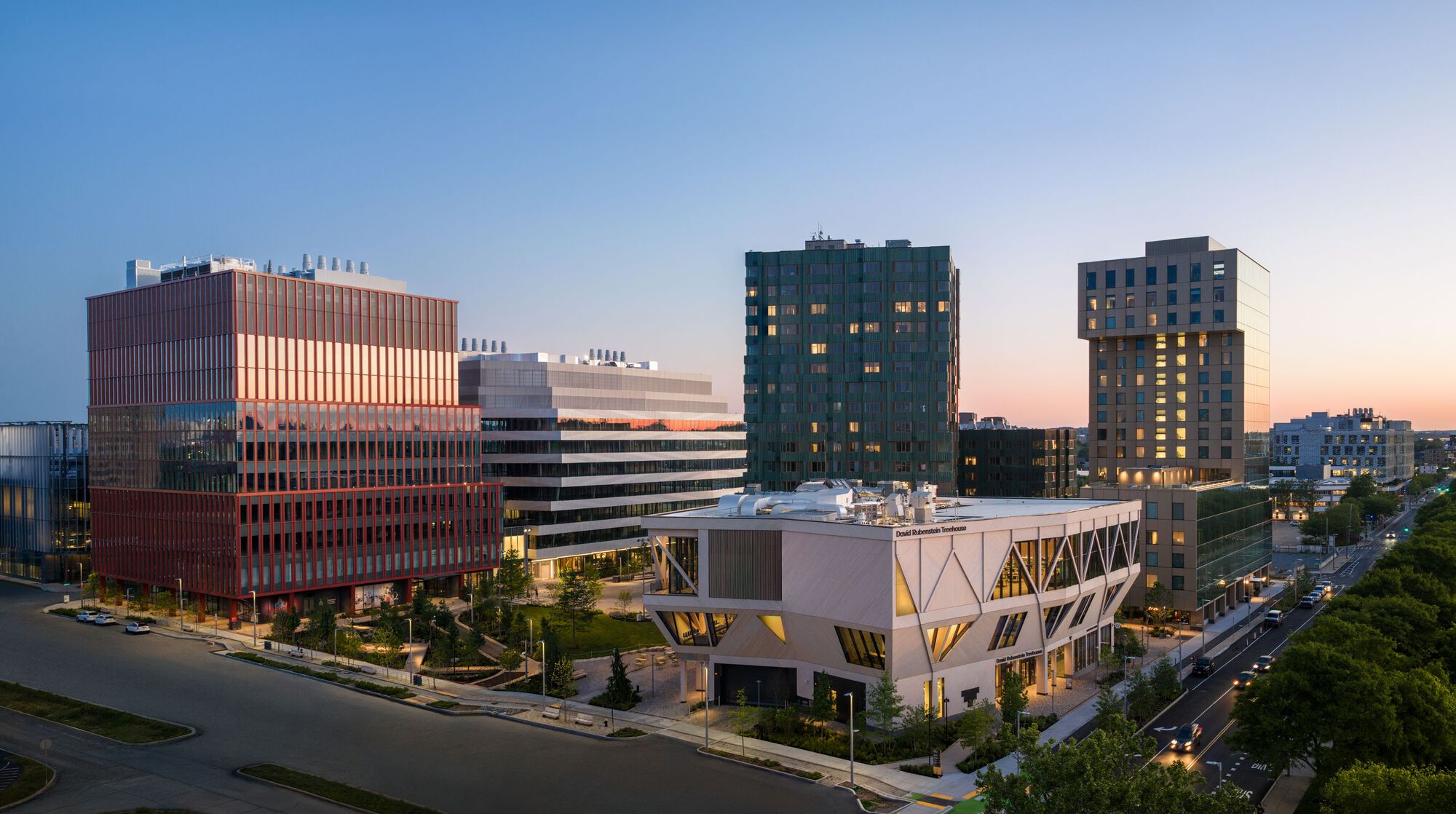 Aerial view of the mixed-use campus with red-paneled facade and white angular pavilion at dusk