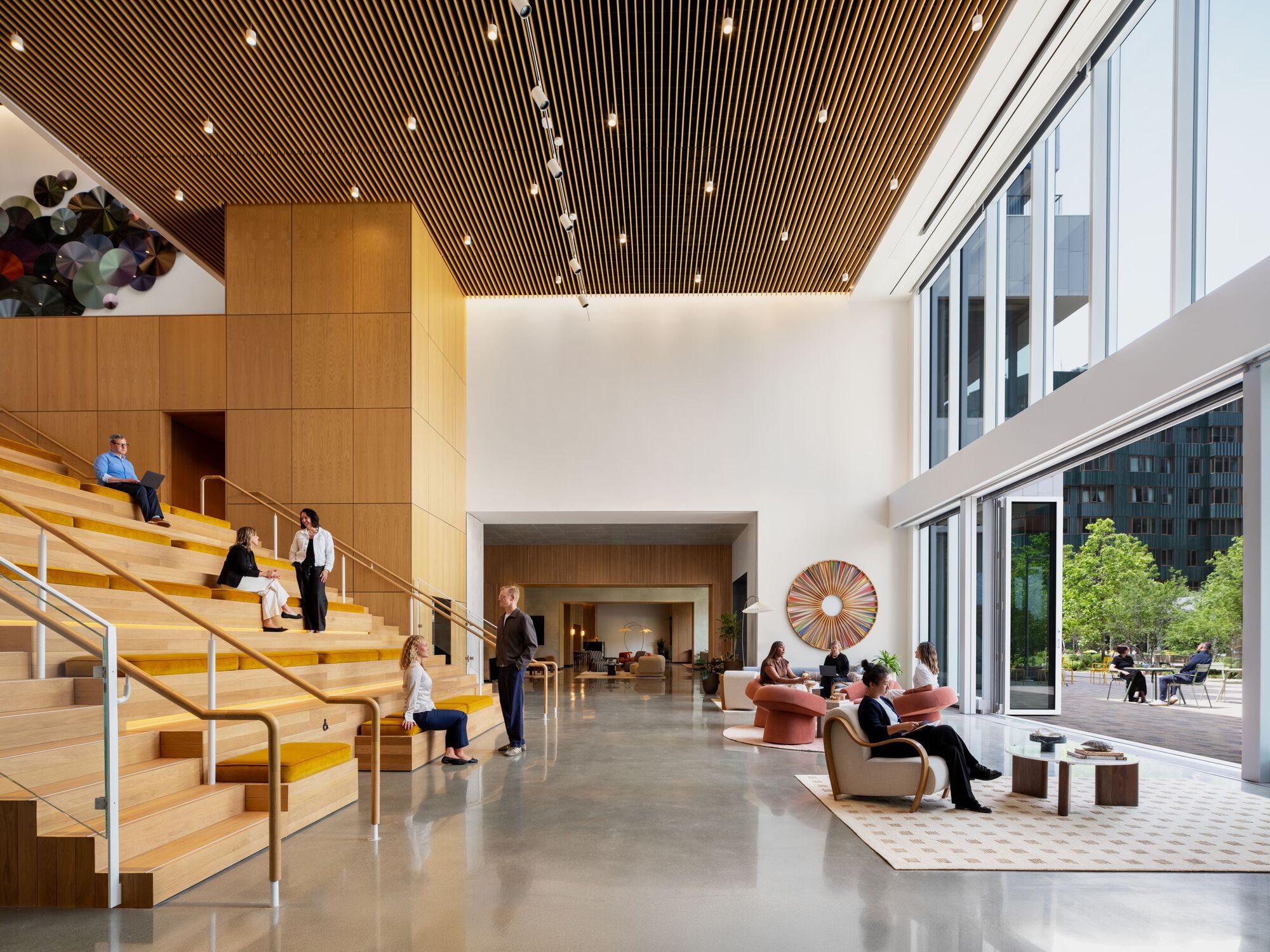 Double-height lobby with wood slat ceiling, tiered seating stair, and people gathering on polished concrete