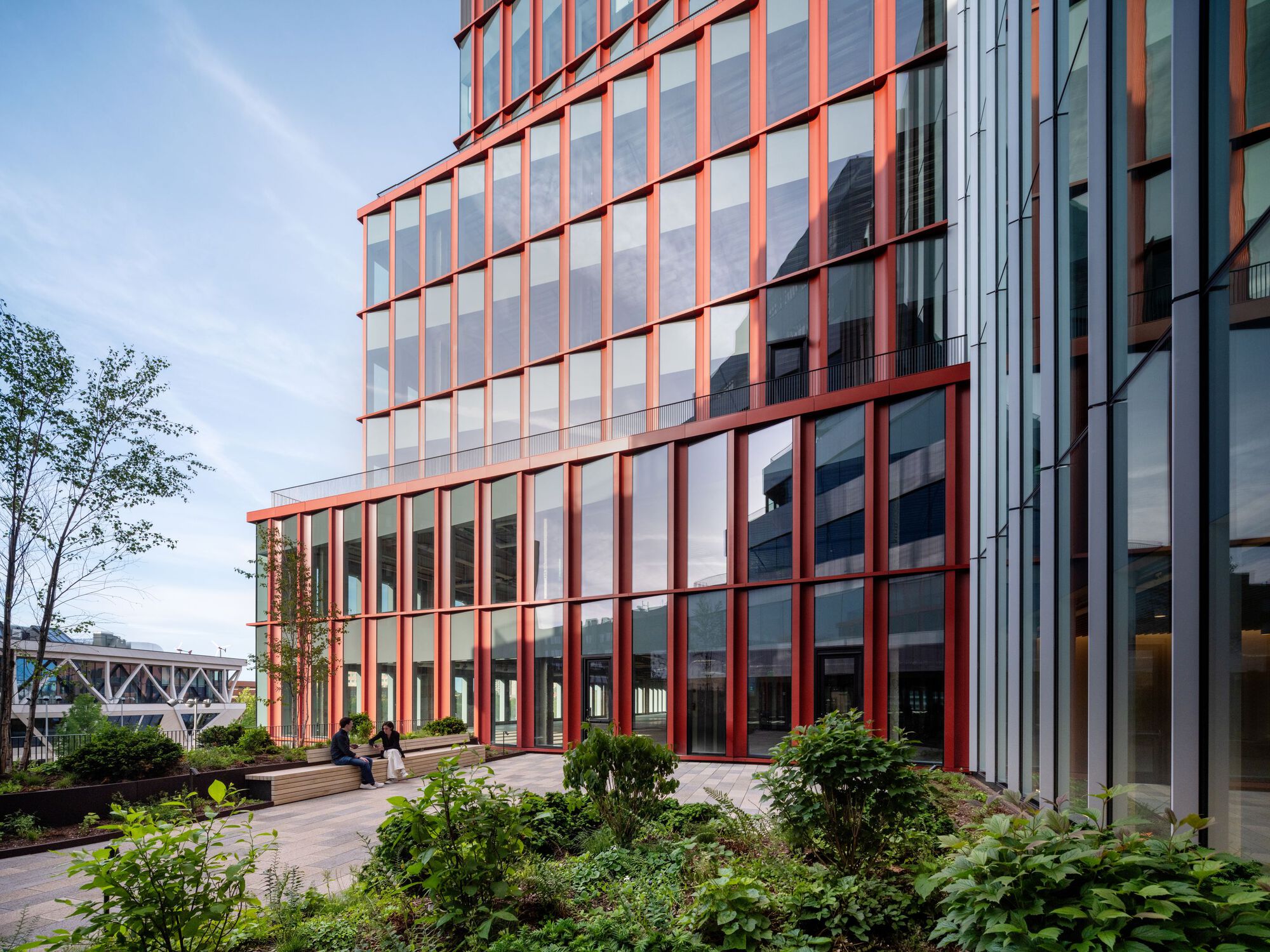 Red steel frame facade over glazed curtain wall with garden beds and walking figures