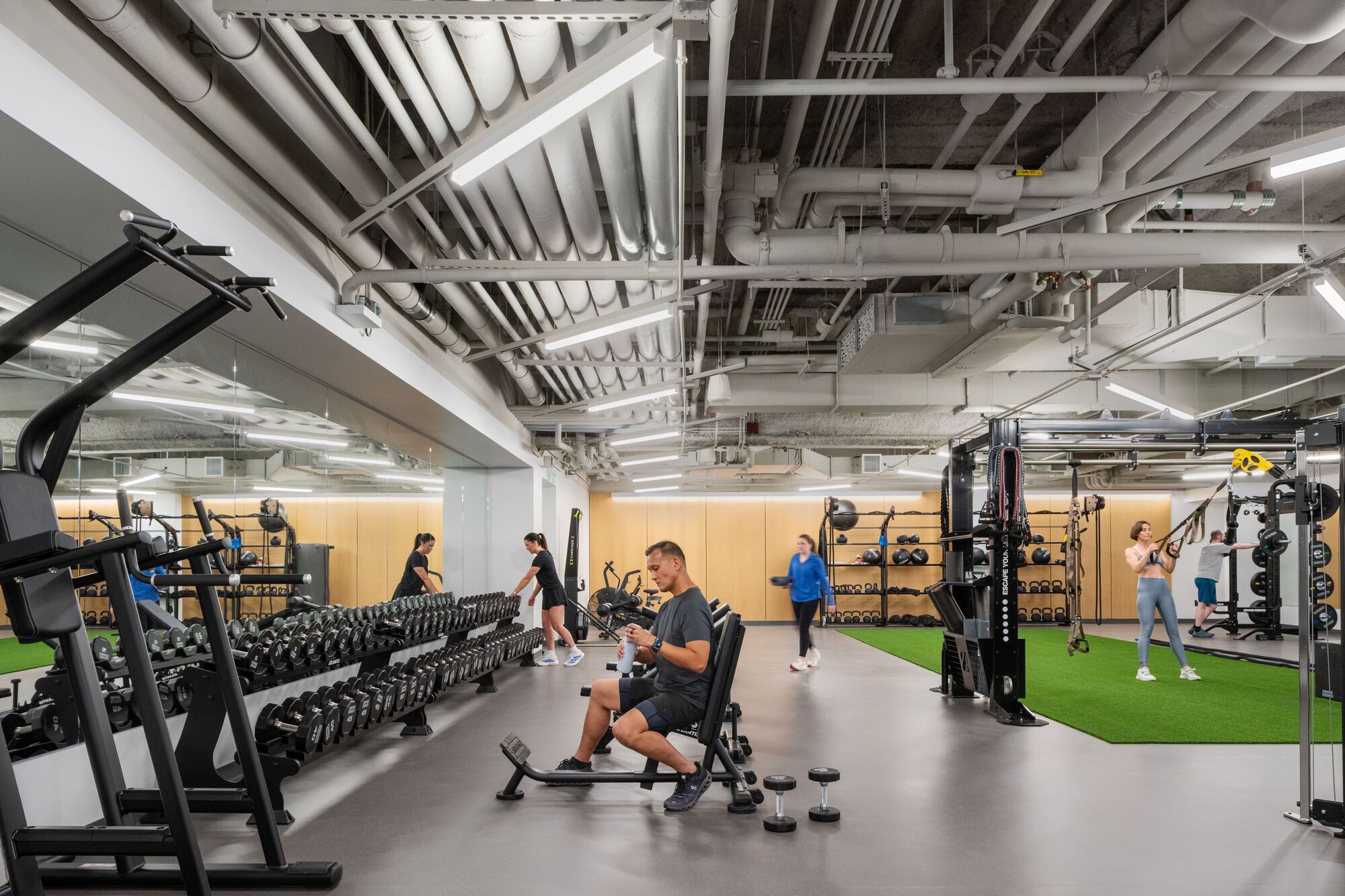 Fitness center with exposed ceiling ducts, white structural beams, and people using free weights