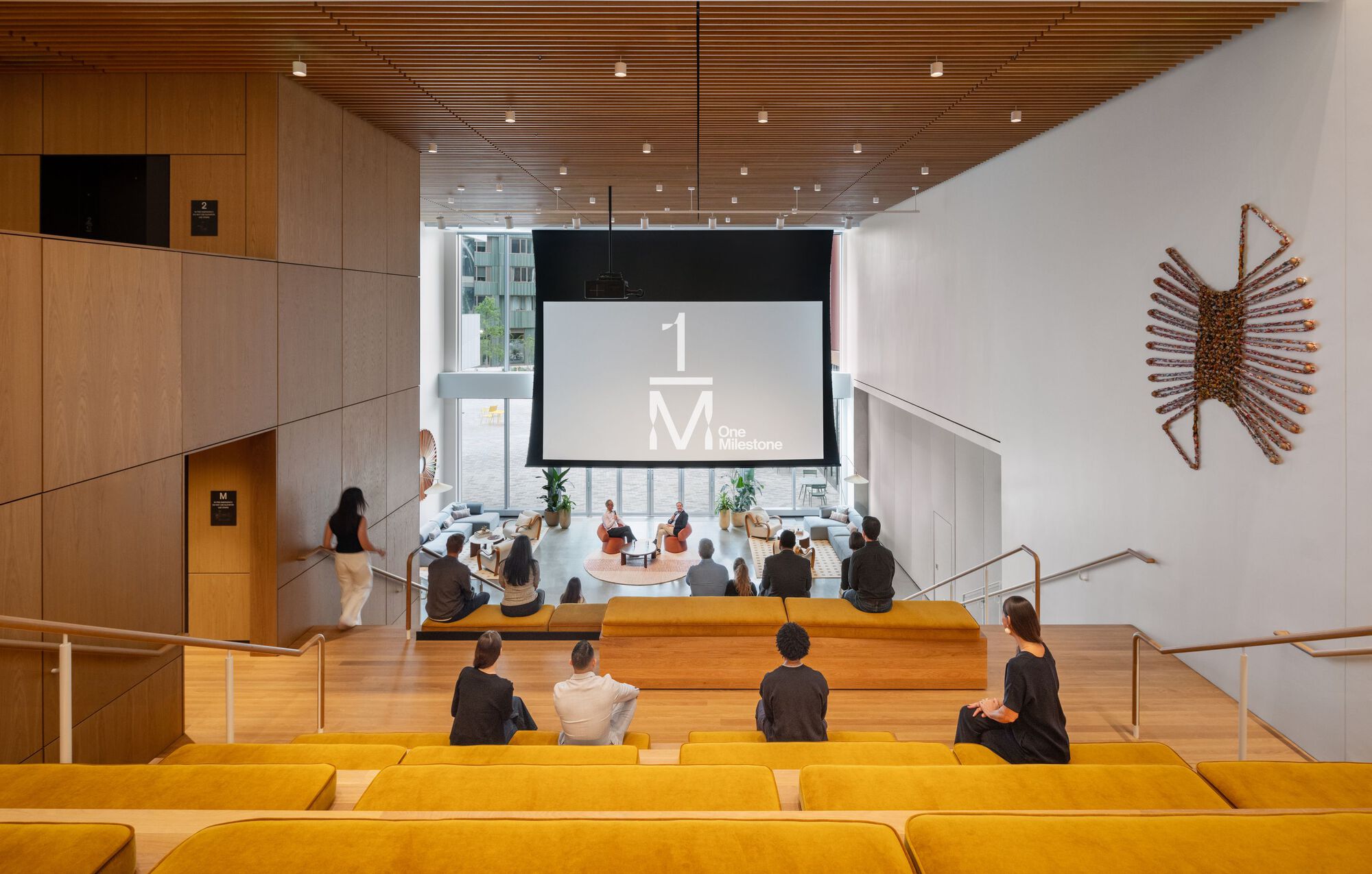 Tiered seating area with yellow cushions facing a projection screen and wall-mounted sculptural art piece