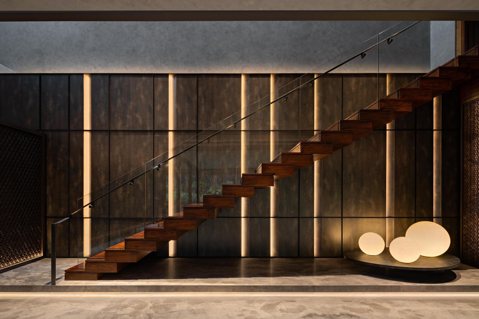 Floating timber staircase with glass balustrade against backlit vertical wood slat wall at dusk