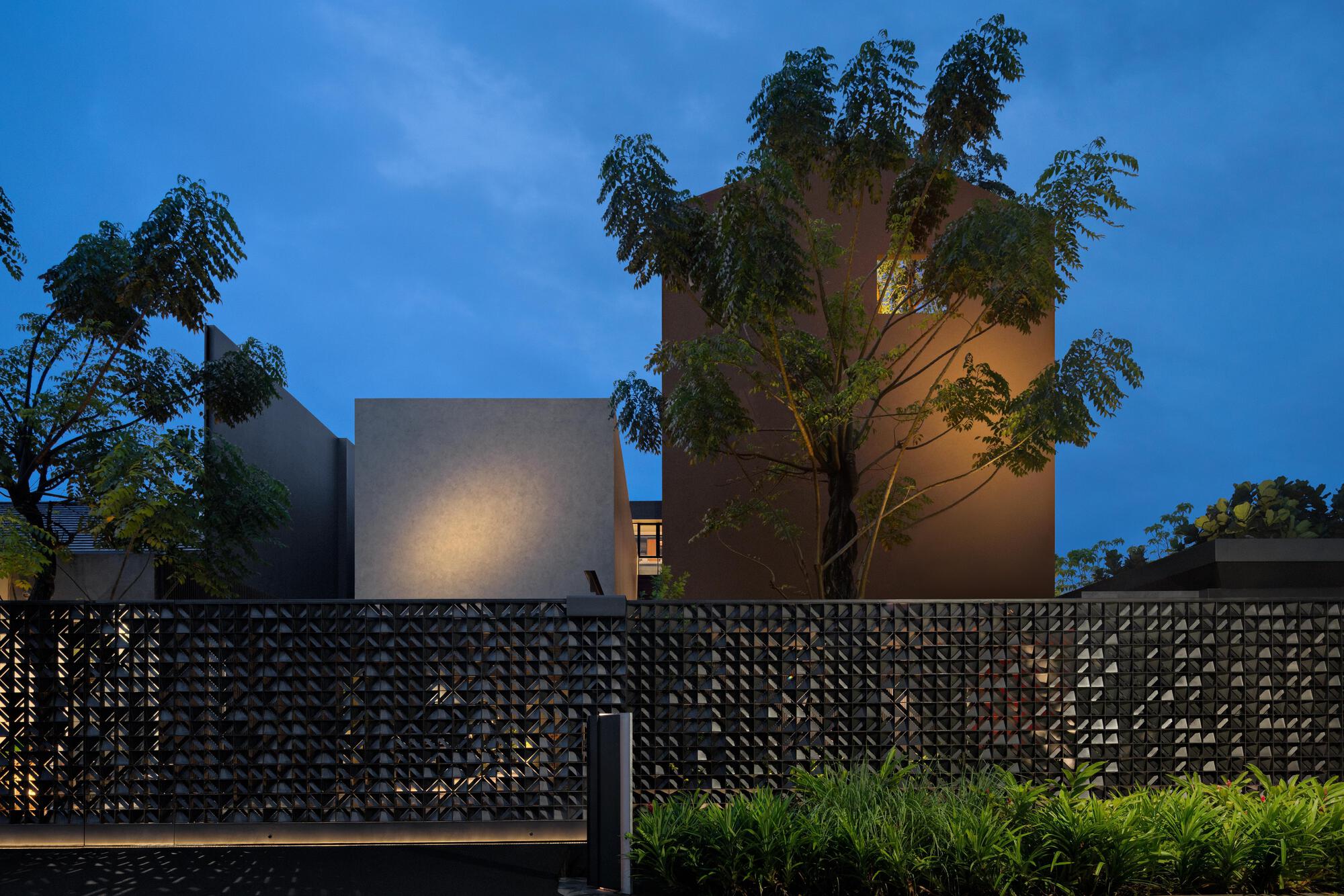 Street view showing stacked volumes behind a perforated black metal fence and leafy tree at twilight