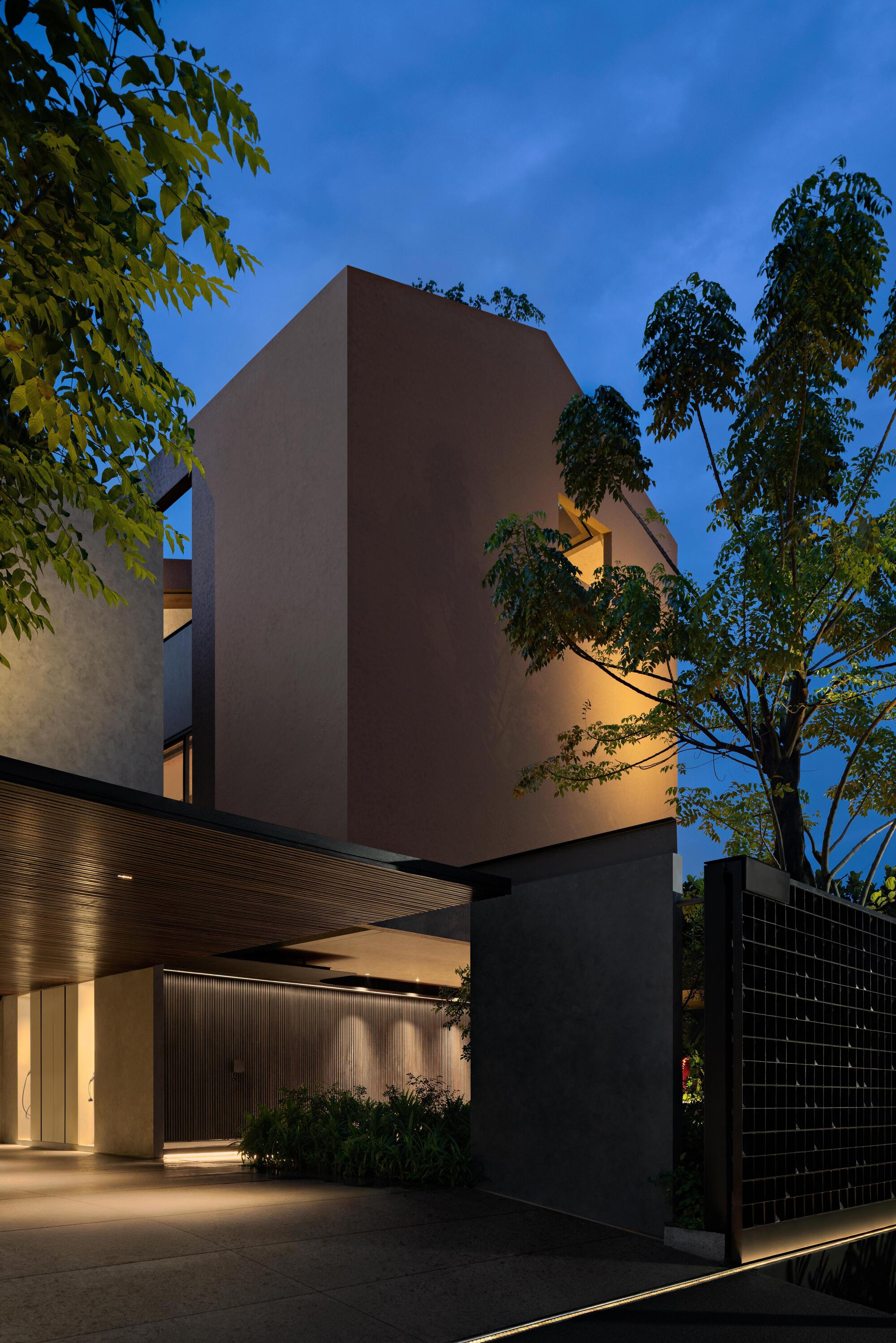 Front entry with illuminated timber-clad carport and tall pink-painted volume framed by trees at dusk