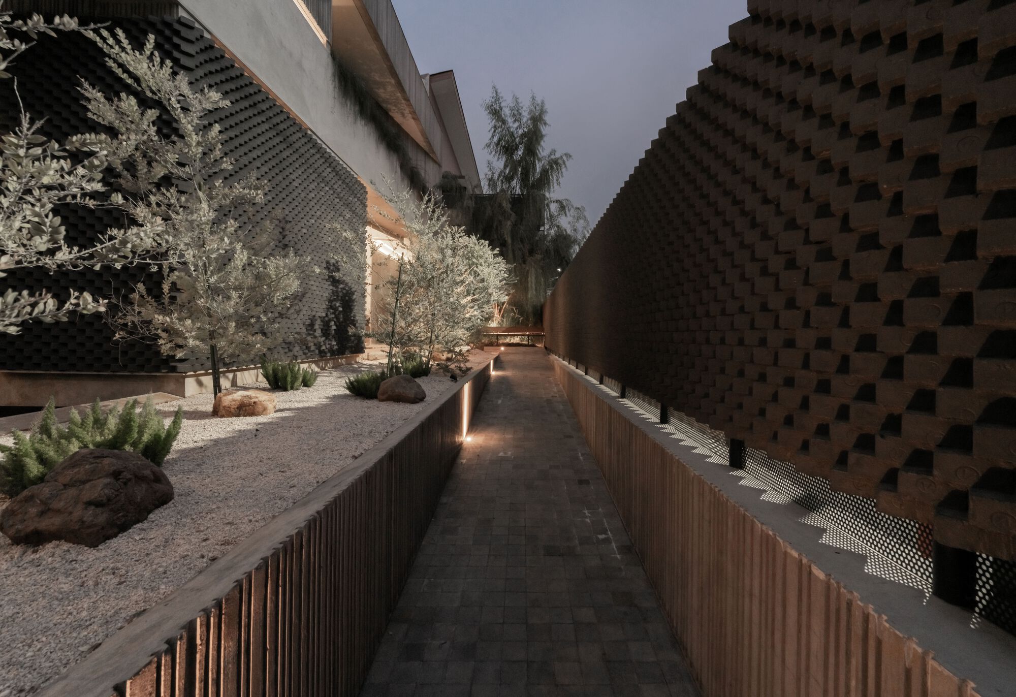 Elevated walkway through courtyard garden with gravel beds, olive trees and perforated brick walls at dusk