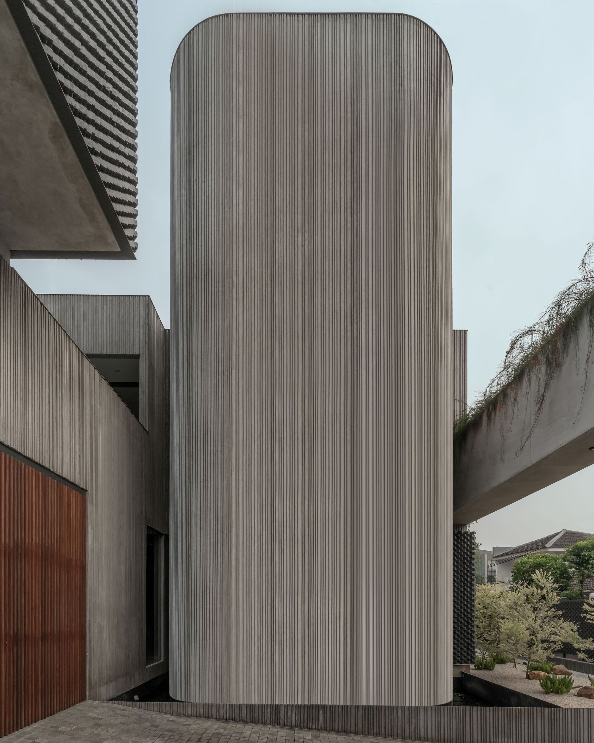 Vertical timber-clad cylinder rising between concrete bridge volume and perforated screen facade under overcast sky
