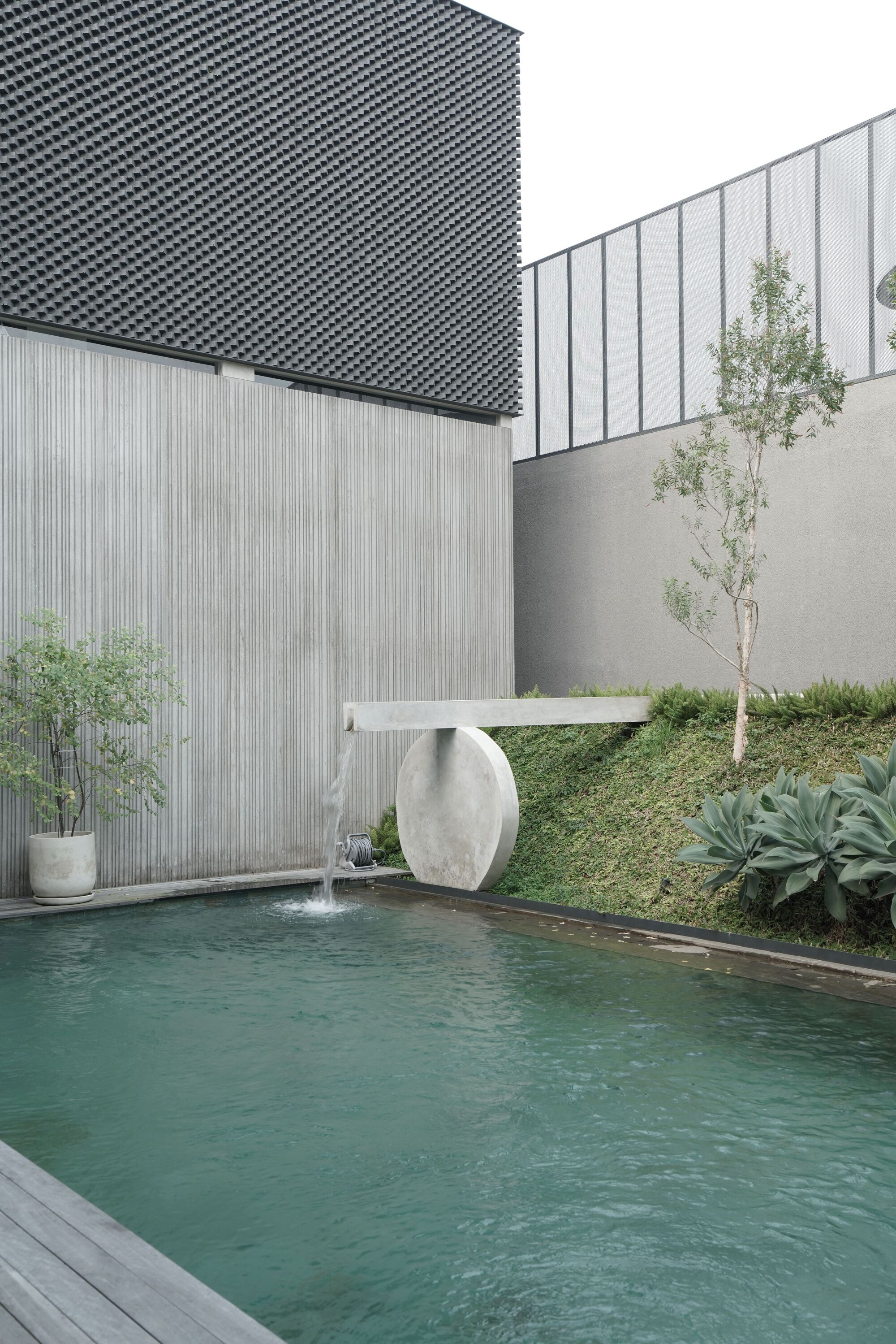 Pool with oval stone fountain sculpture beside planted slope with young trees and perforated screen volume