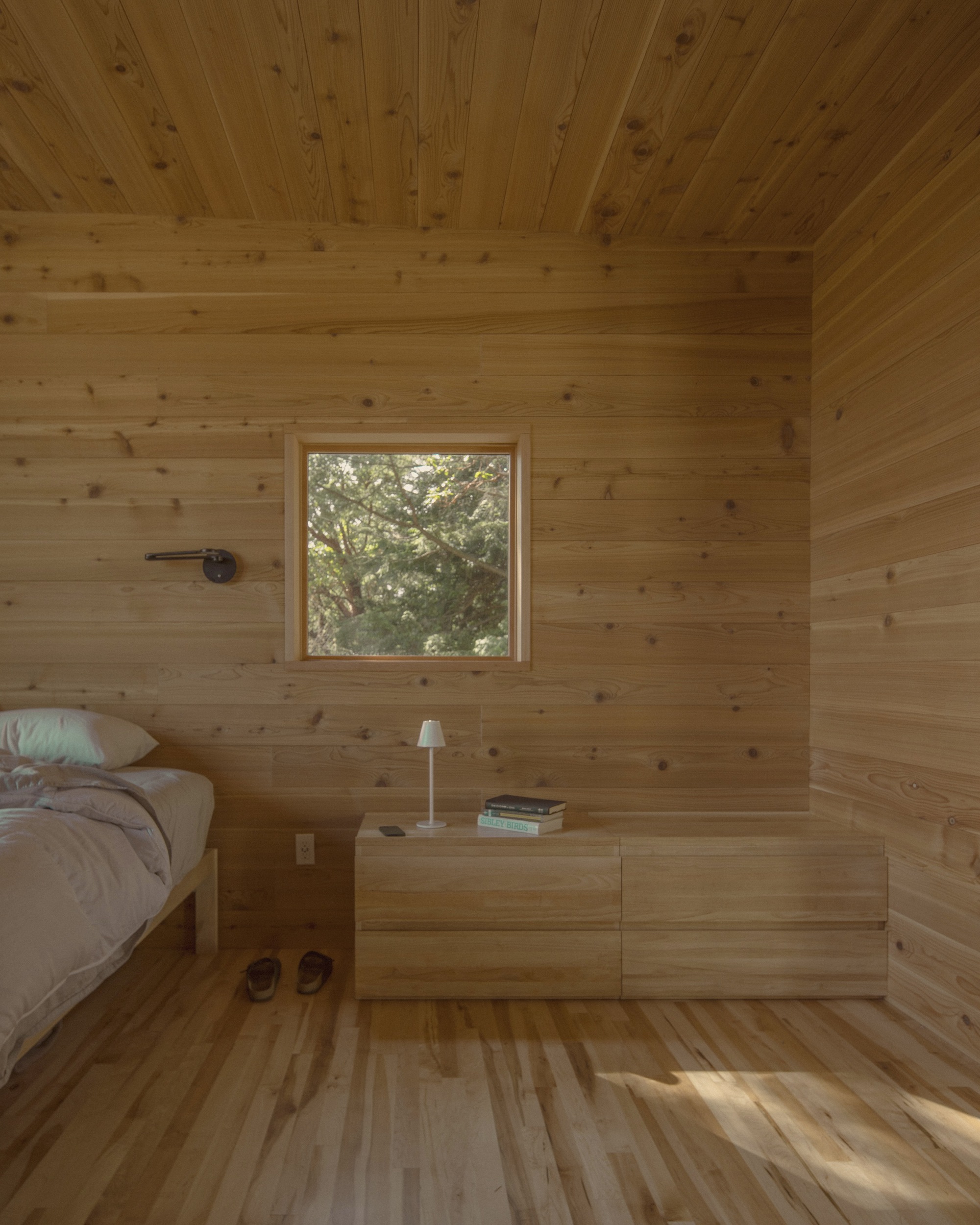 Timber-lined bedroom with square window framing trees and built-in dresser catching afternoon sunlight