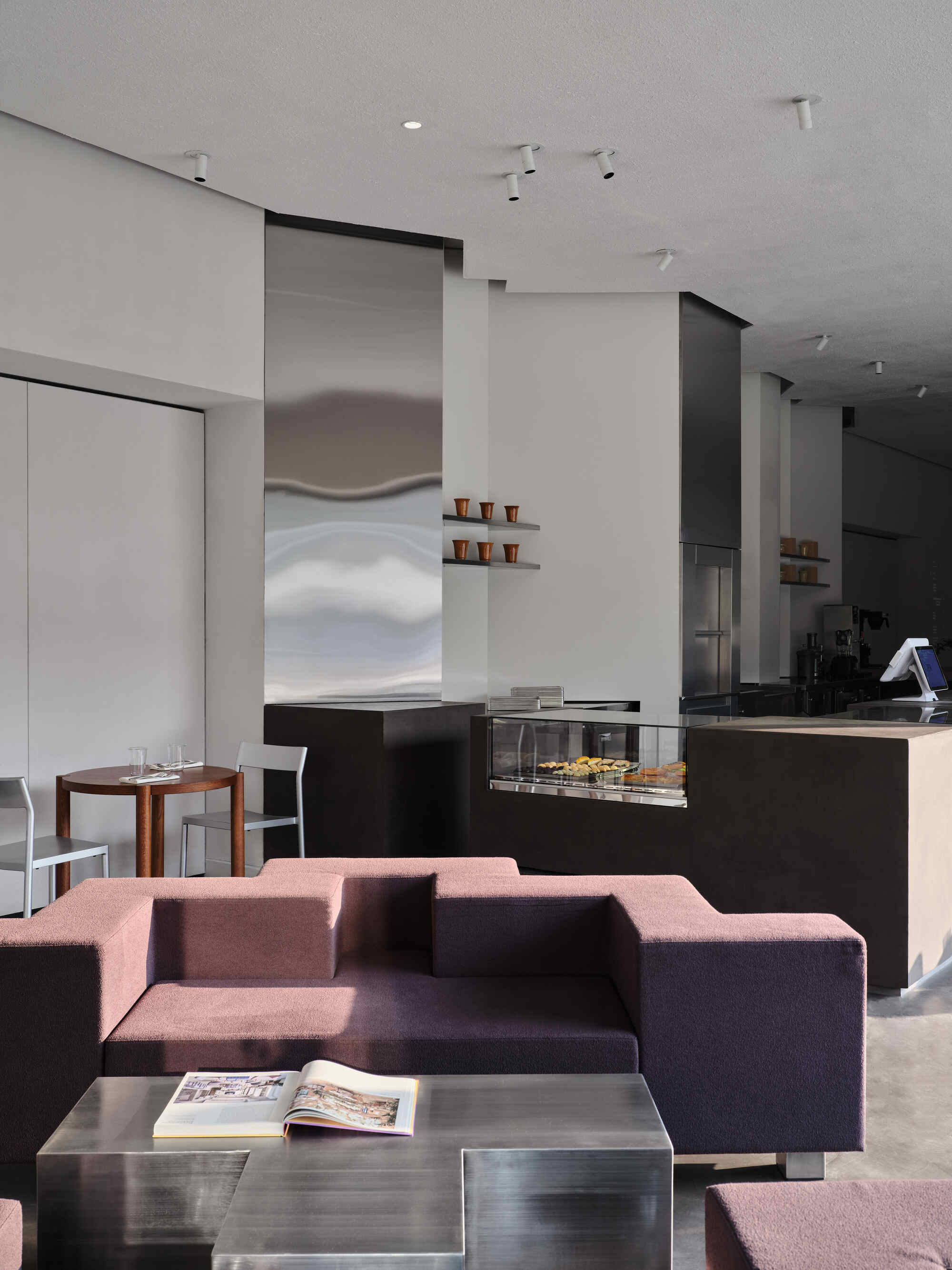 Open-plan space showing the lounge with burgundy seating foreground and black service counter beyond stainless steel column