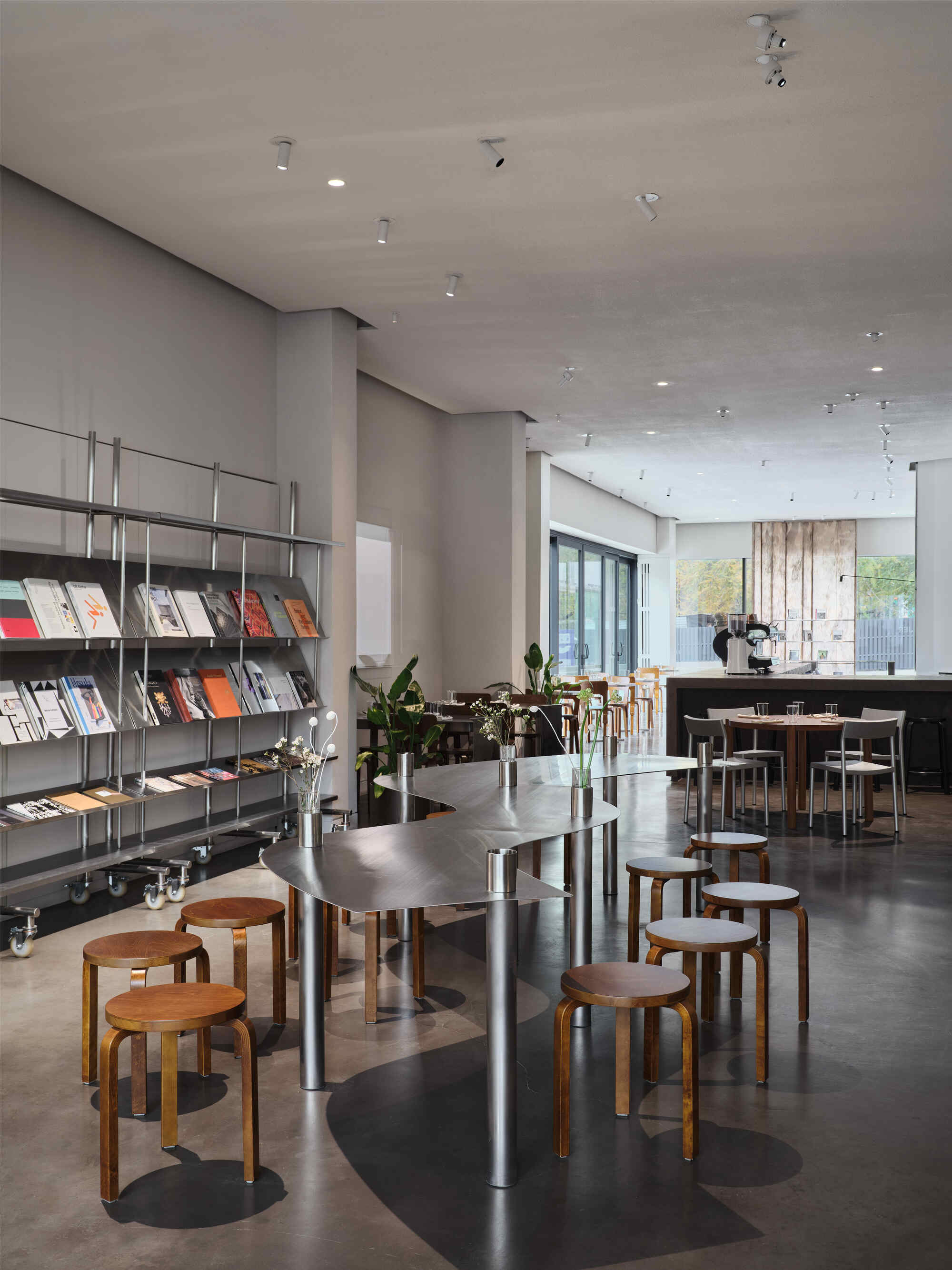 Open cafe space with magazine shelving, timber stools, and daylight through glazed doors beyond potted plants