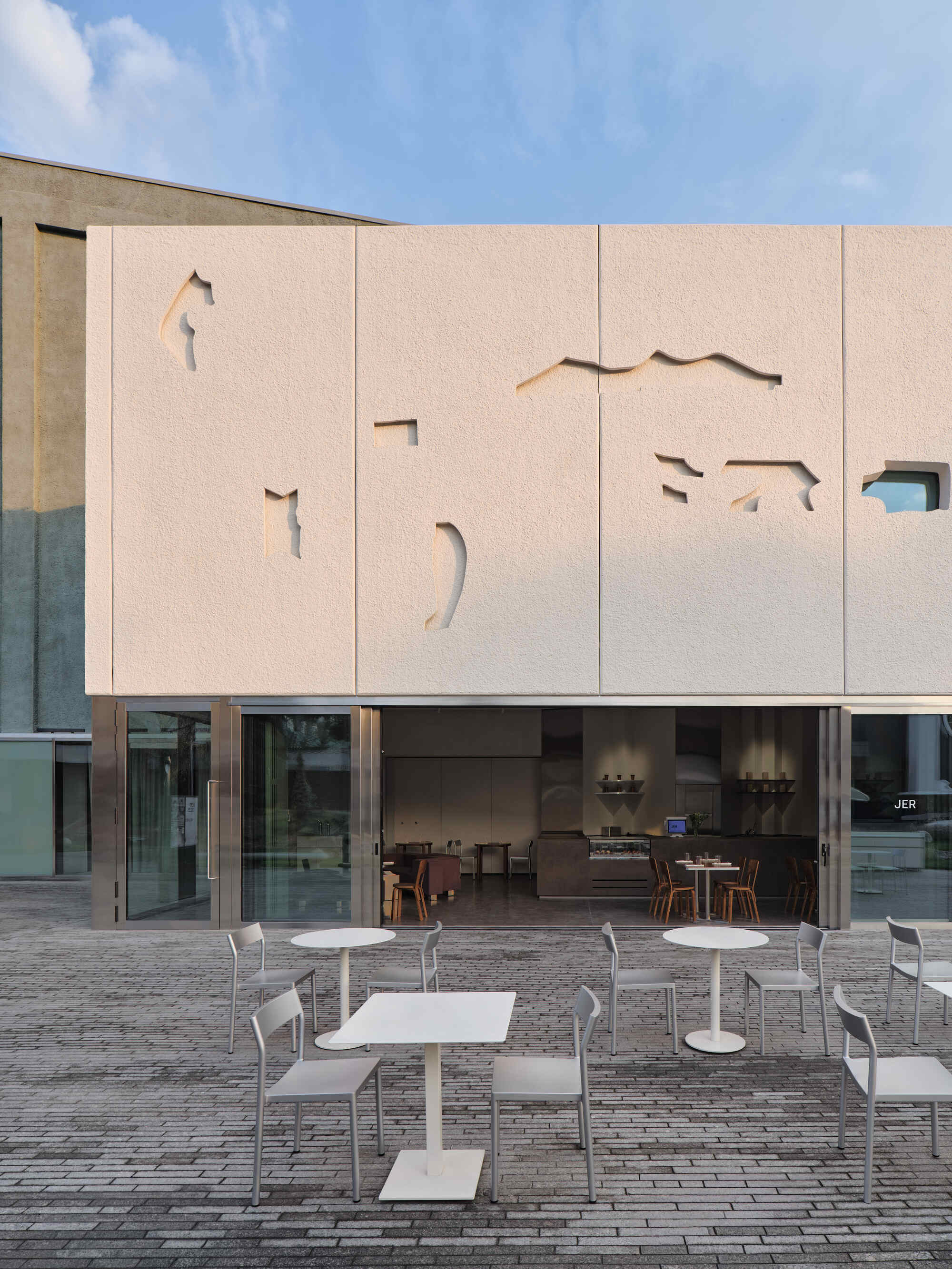Pale facade with cutout animal shapes above a glass-walled cafe terrace with white outdoor furniture