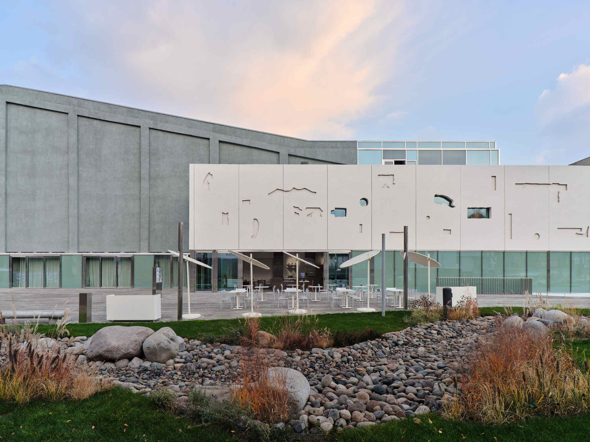 Cantilevered facade with organic cutouts above a glazed base, adjacent to a landscaped rock garden at dusk