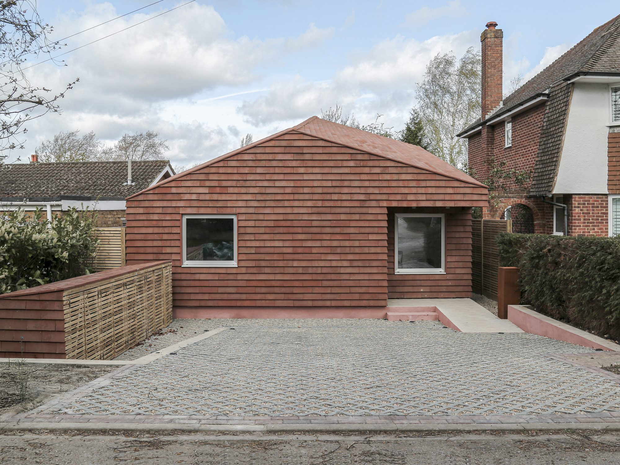 Front view of the terracotta tile-clad volume with two square windows and permeable paving under cloudy skies