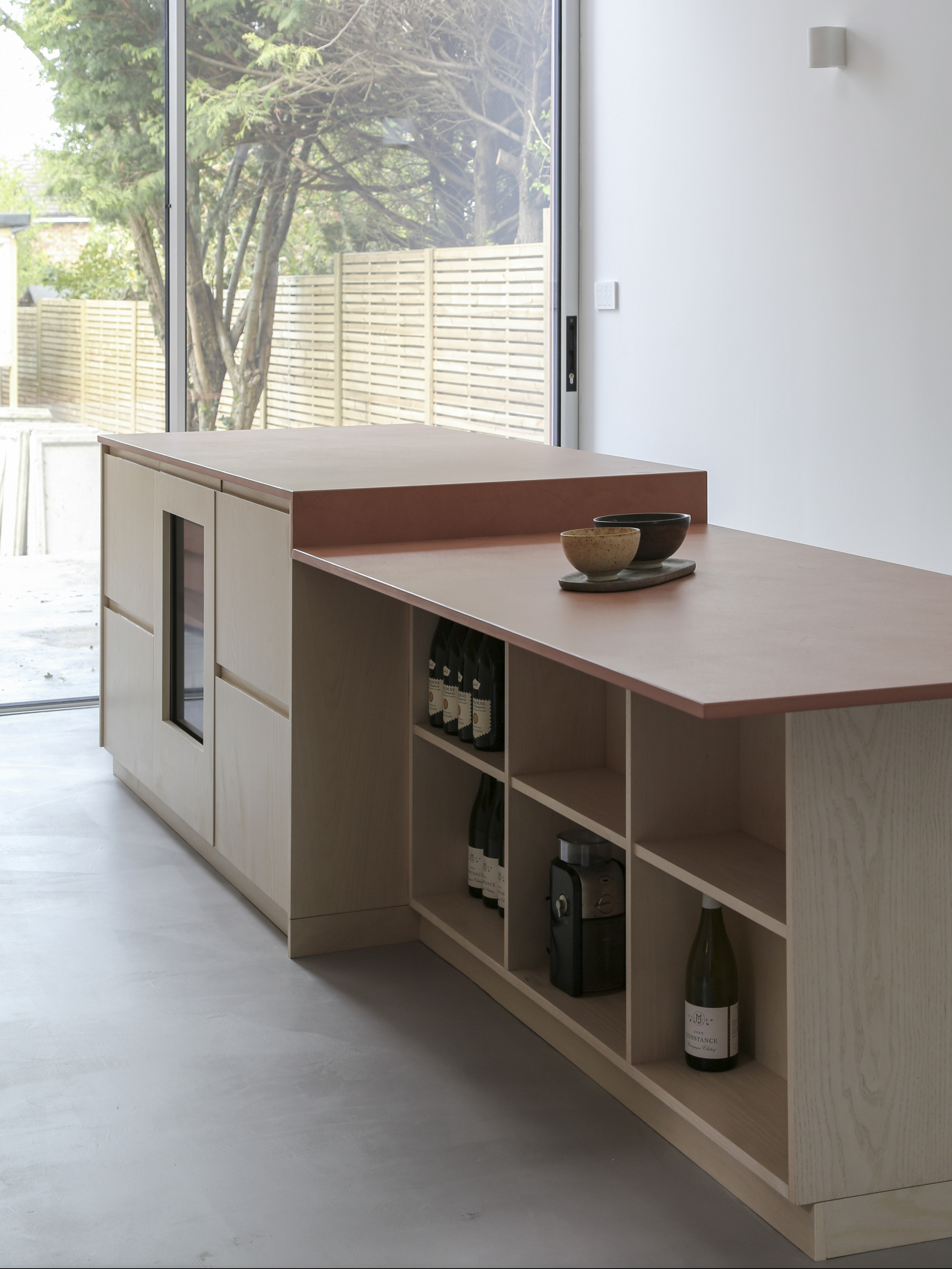 Kitchen island with open shelving for wine storage and view to horizontal slatted fence beyond glazing