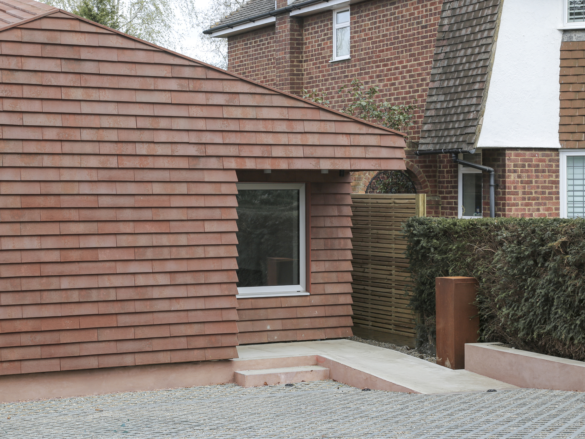Curved timber-clad extension wall meeting a paved courtyard entrance with trimmed hedge beyond