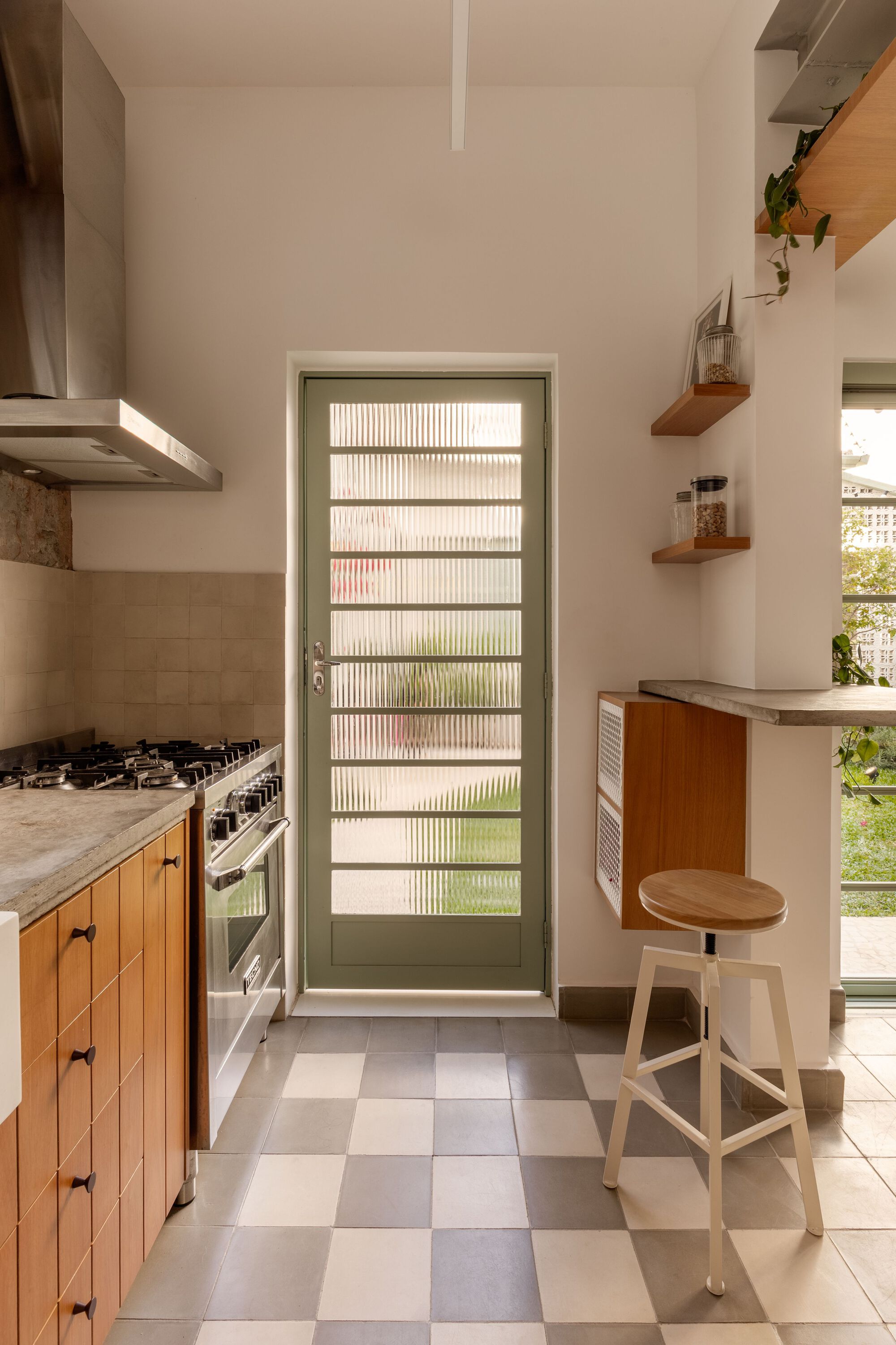 Narrow kitchen passage with checkered floor, timber cabinetry, green-framed glass door, and floating timber shelves
