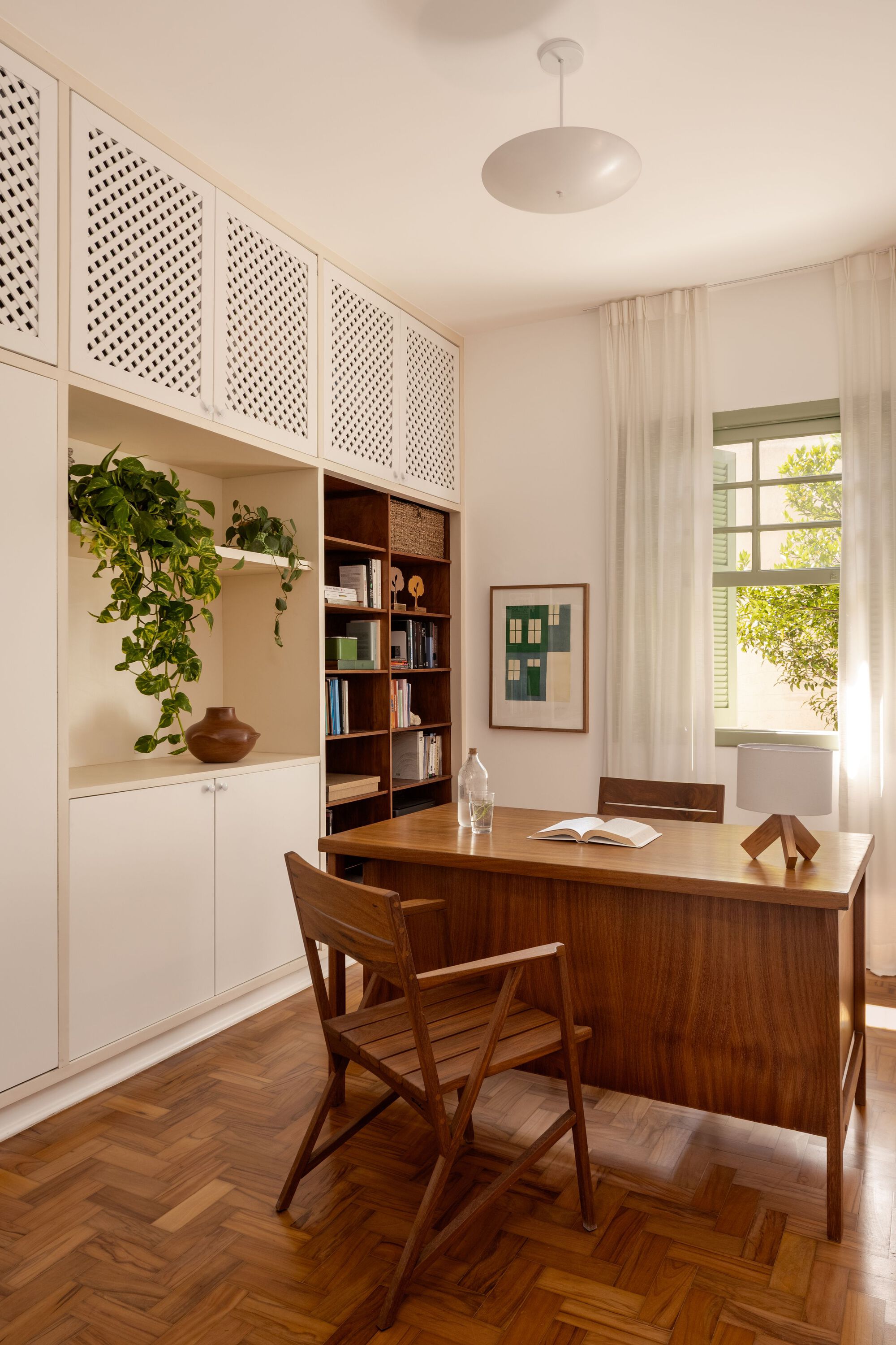 Home office with walnut desk, perforated upper cabinets, herringbone wood floor and potted trailing plants