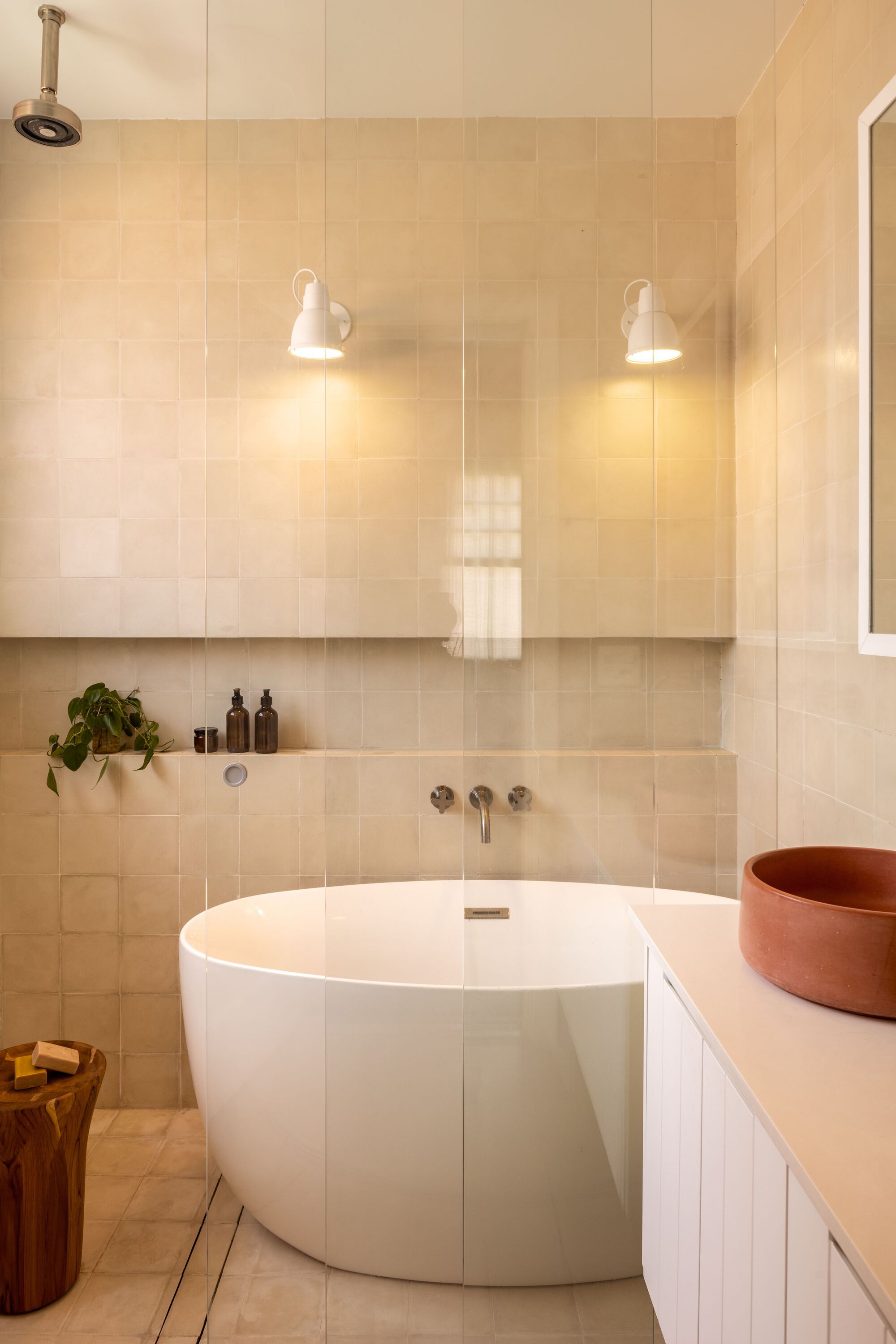 Bathroom with freestanding white bathtub beneath cream tile walls and paired wall-mounted sconces