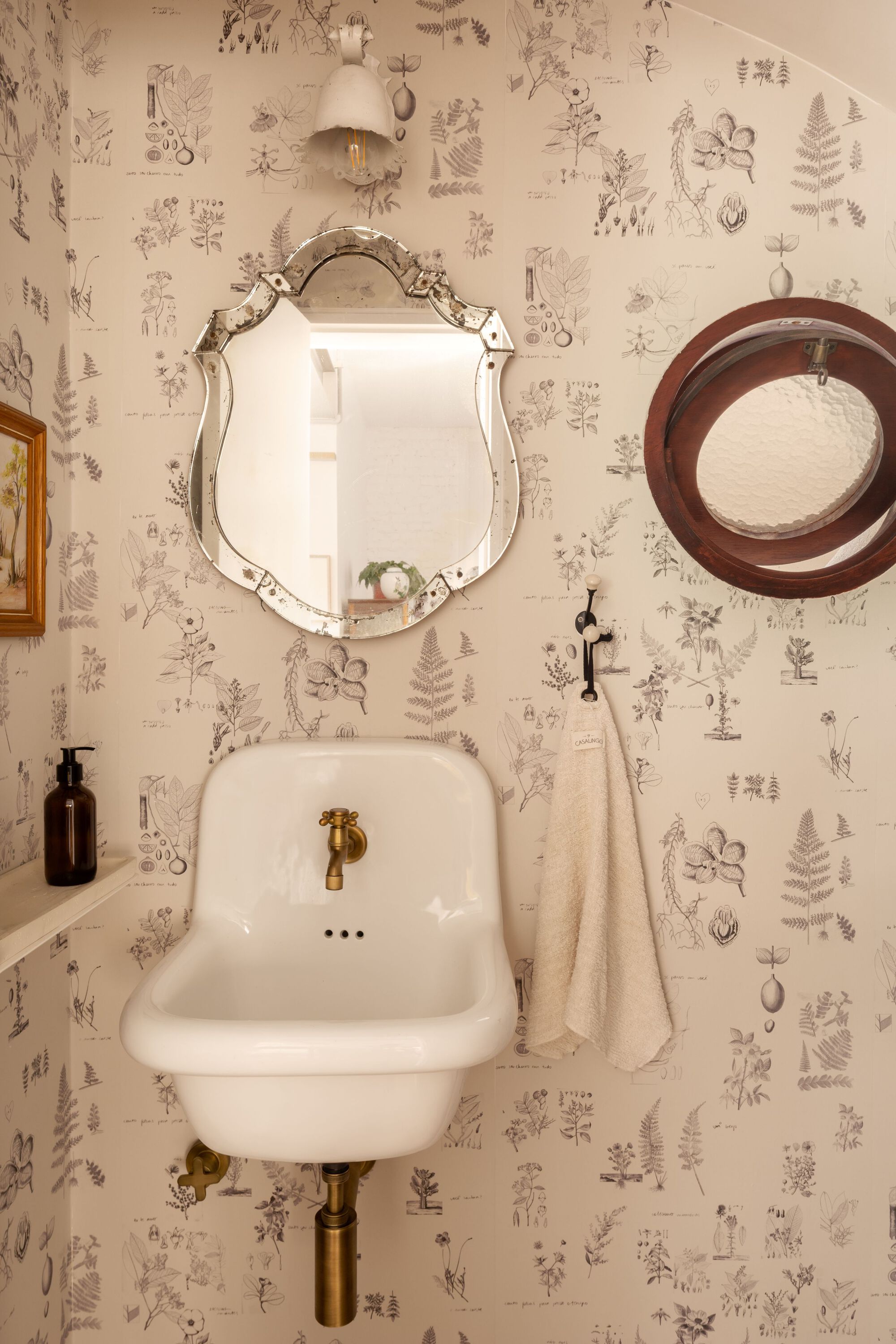Wall-mounted porcelain sink with brass fixtures against botanical patterned wallpaper and ornate mirror