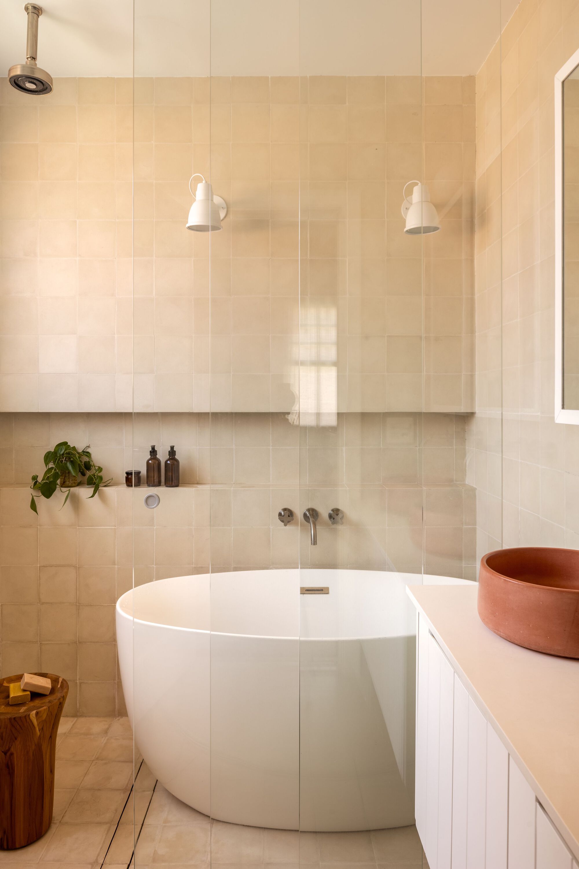 Bathroom with freestanding white tub against pale tile wall and white vanity with terracotta basin