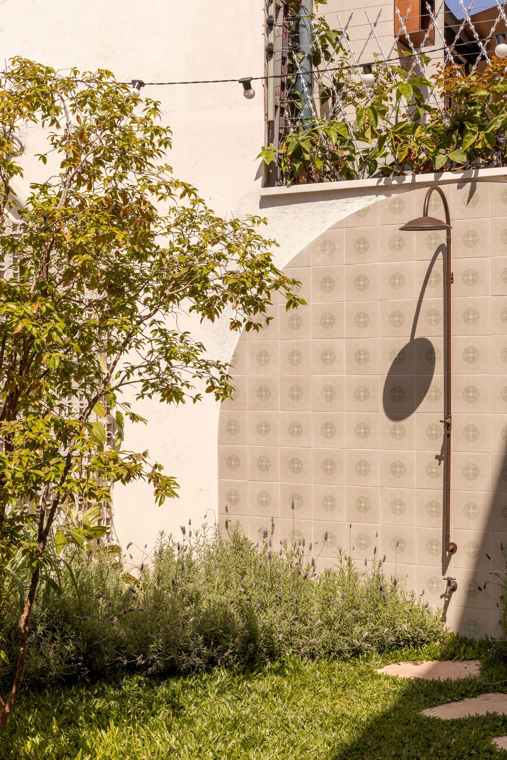 Outdoor shower fixture on patterned tile wall beside planted tree and grass in afternoon sun