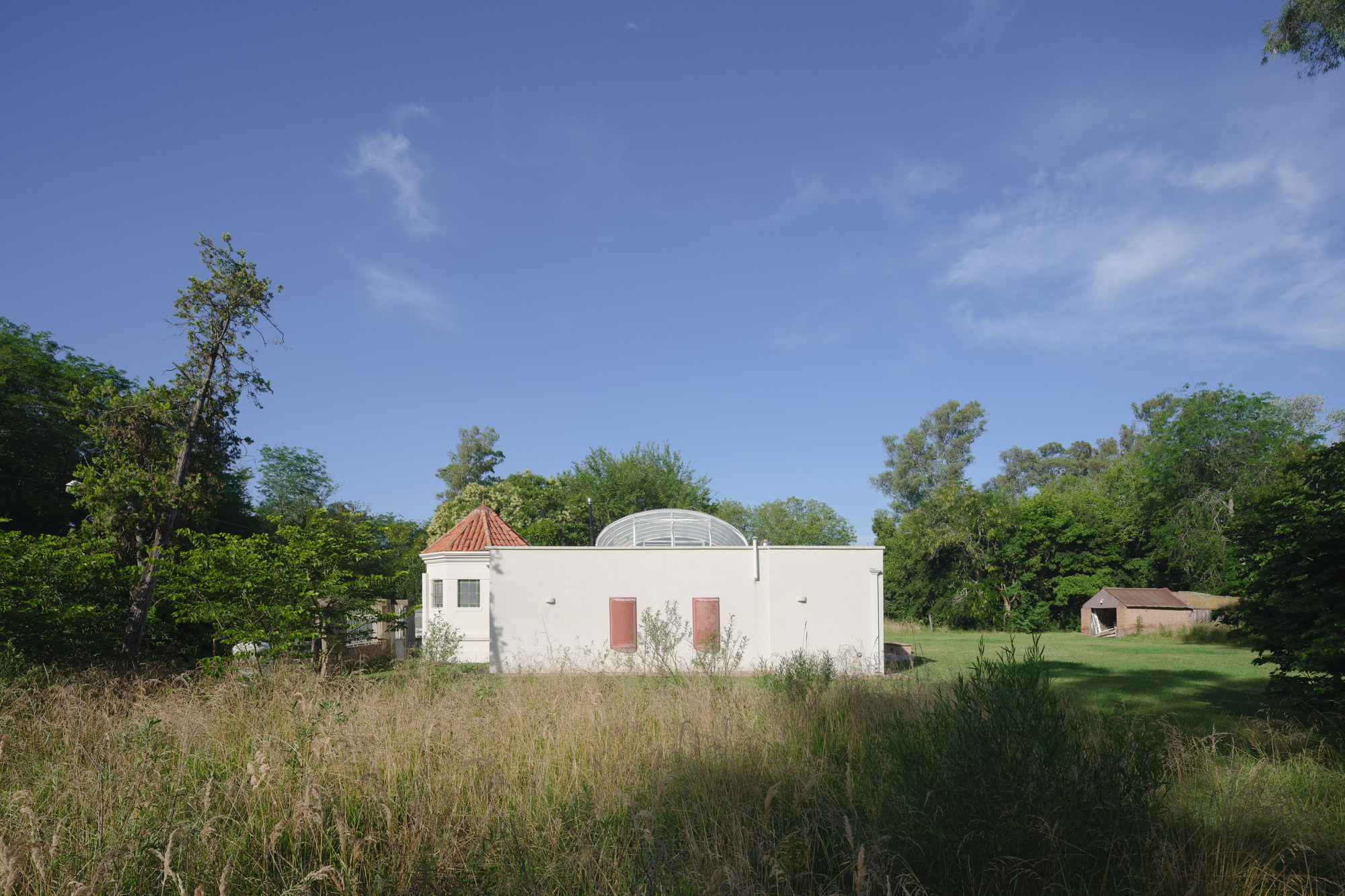 Exterior view of white plastered volume with arched canopy roof surrounded by wildflower meadow and trees