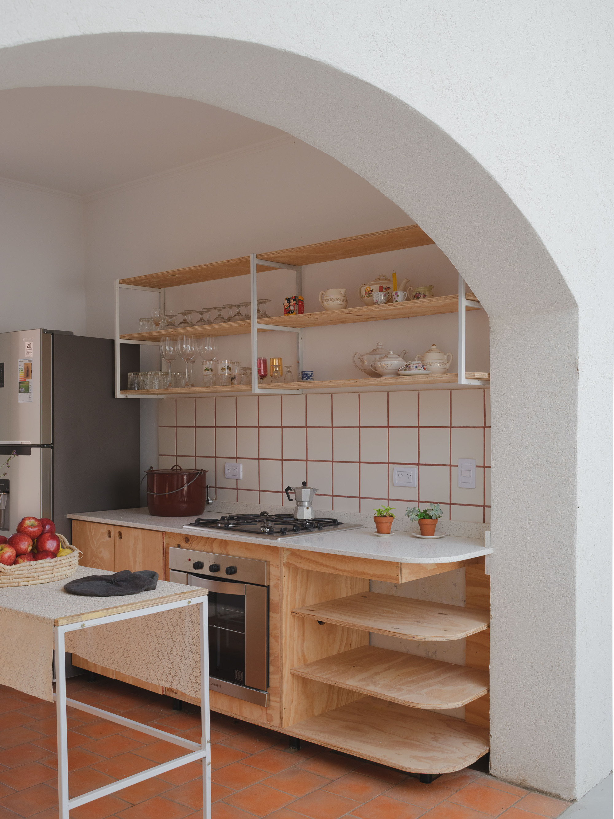 Kitchen niche framed by arched opening with plywood shelving, white tile backsplash and terracotta floor